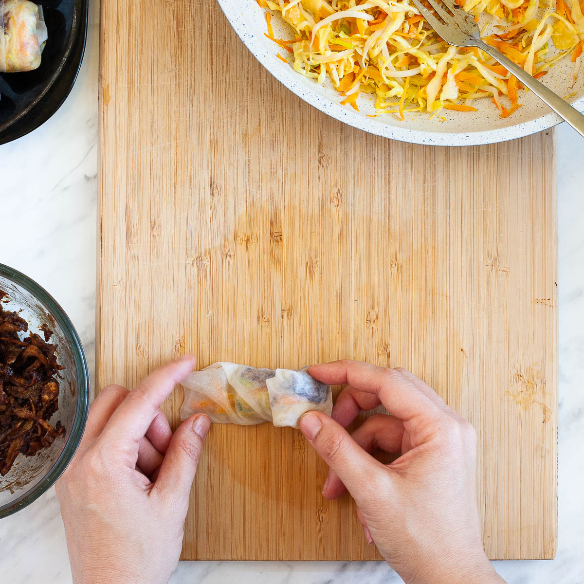 A hand is rolling a rice paper with the filling from sideways to shape a round swirl-like dumpling. It is on a wooden cutting board surrounded by other ingredients in a pan and in a glass bowl.