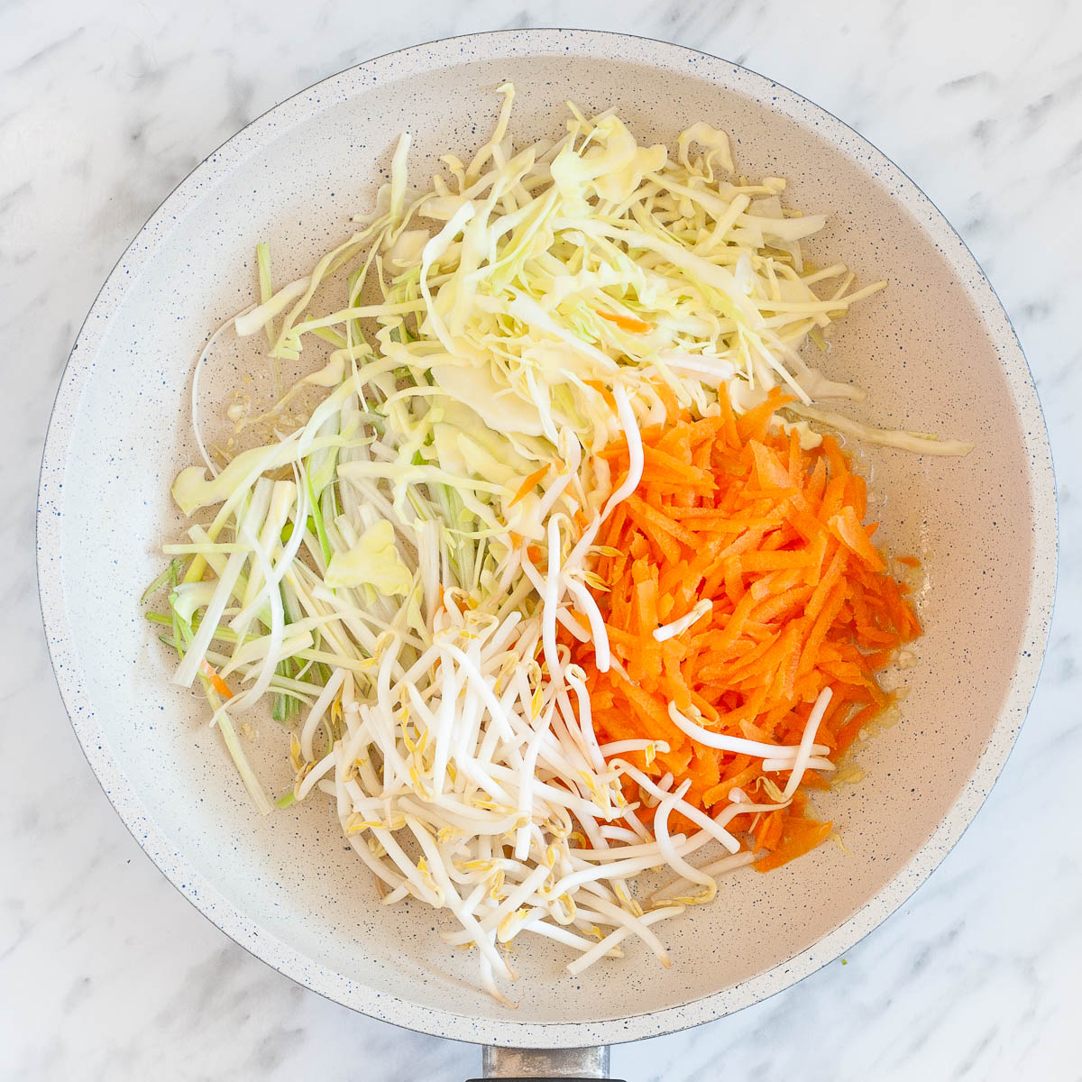 A white frying pan from above full of white and orange shredded veggies like carrots, cabbage, spring onion and sprouts raw before cooking