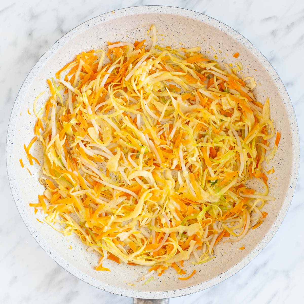 A white frying pan from above full of white and orange shredded veggies like carrots, cabbage, spring onion and sprouts after cooking