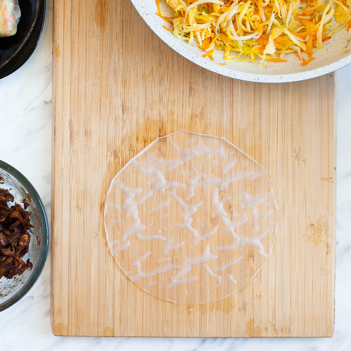 Wet rice paper on a wooden cutting board, surrounded by the ingredients for the dumplings in a pan and in a glass bowl.
