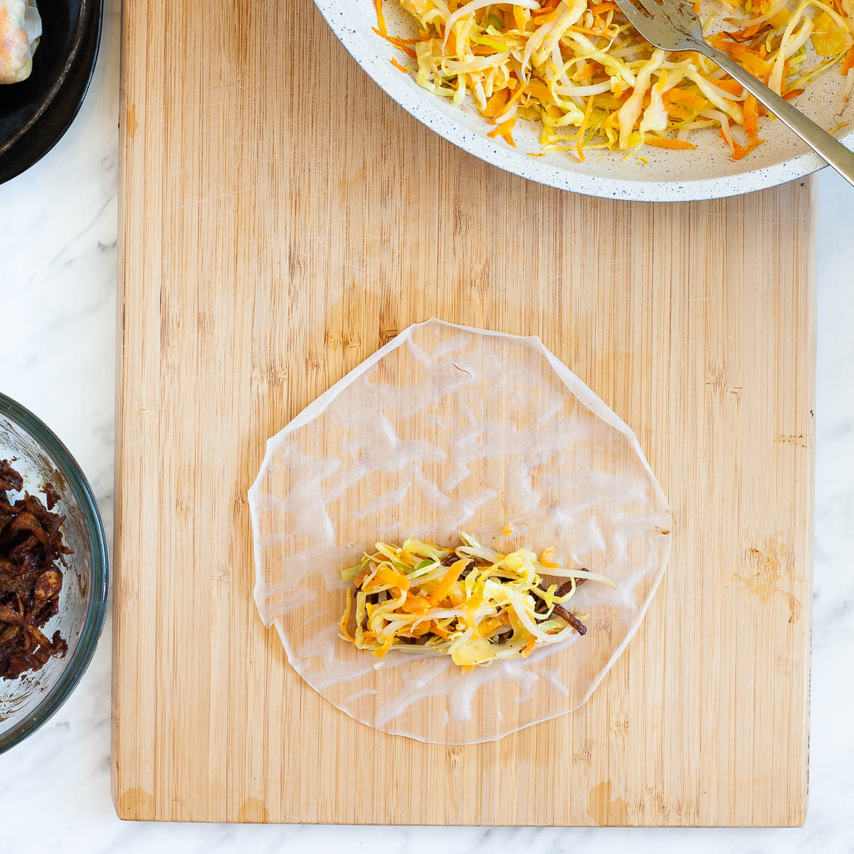 Wet rice paper with some colorful filling on it placed on a wooden cutting board, surrounded by the ingredients for the dumplings in a pan and in a glass bowl.