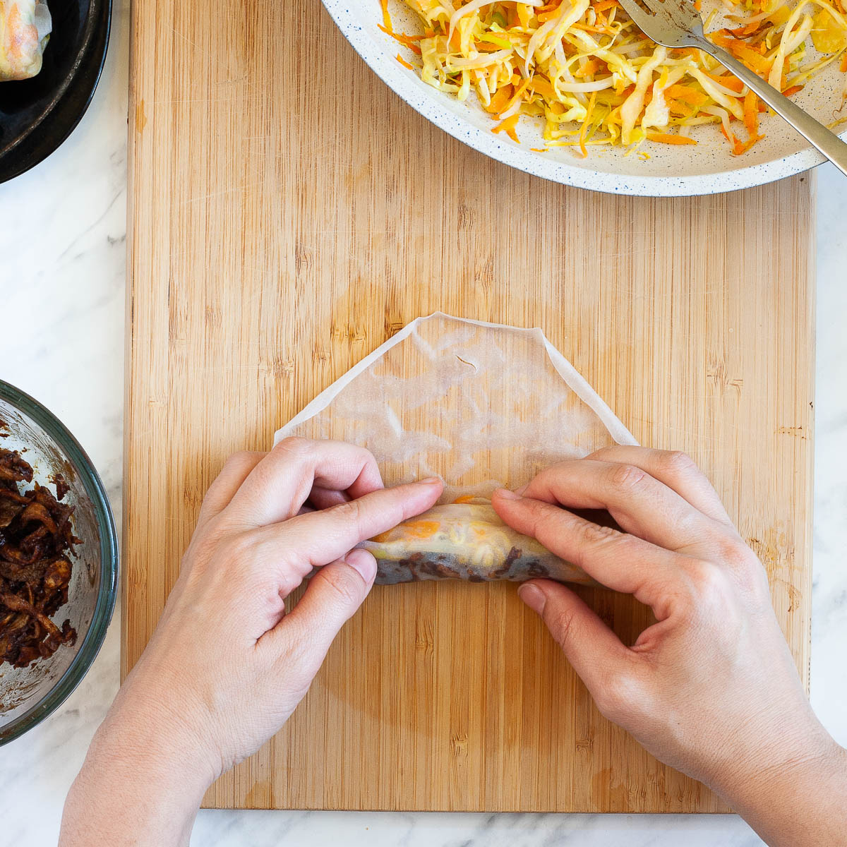 A hand is rolling a rice paper with the filling from bottom to top in a shape of a rod. It is on a wooden cutting board surrounded by other ingredients in a pan and in a glass bowl.