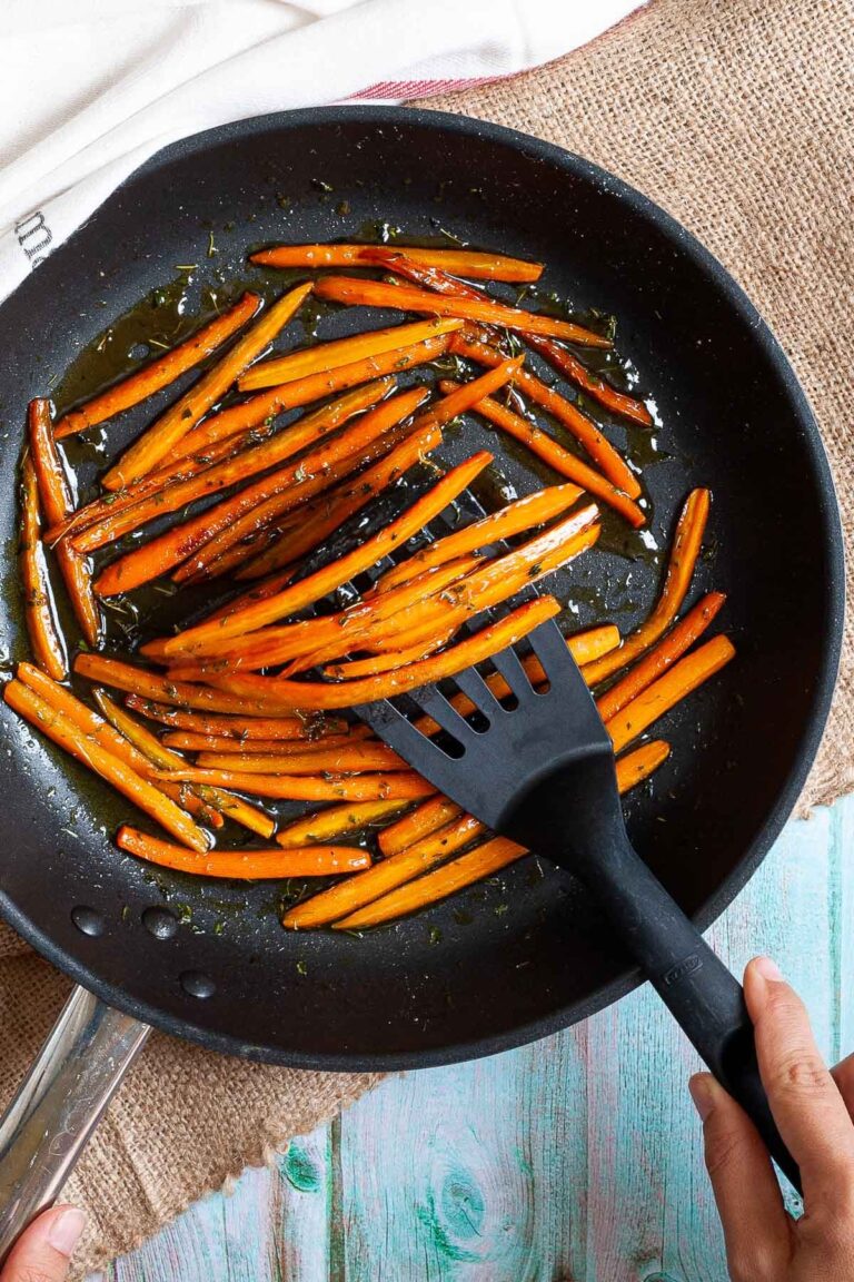 Frying pan with matchstick carrots in a glaze. A hand is holding a slotted turner and taking some from the pan.