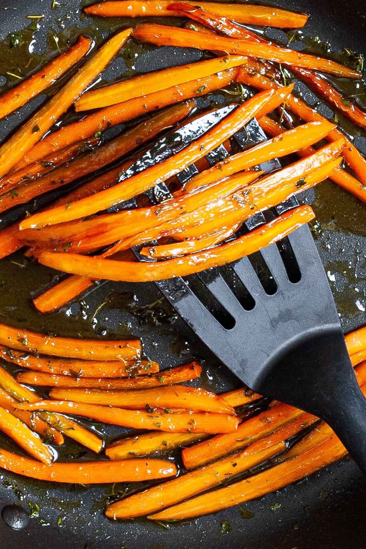 Frying pan with matchstick carrots in a glaze. A hand is holding a slotted turner and taking some from the pan.