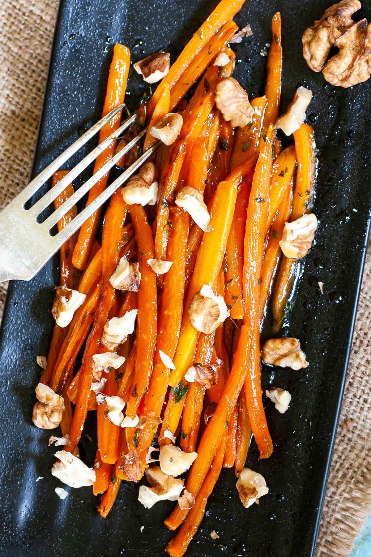 Black rectangular plate with matchstick carrots in glaze and walnut pieces. A hand is holding a fork and taking some from the top.