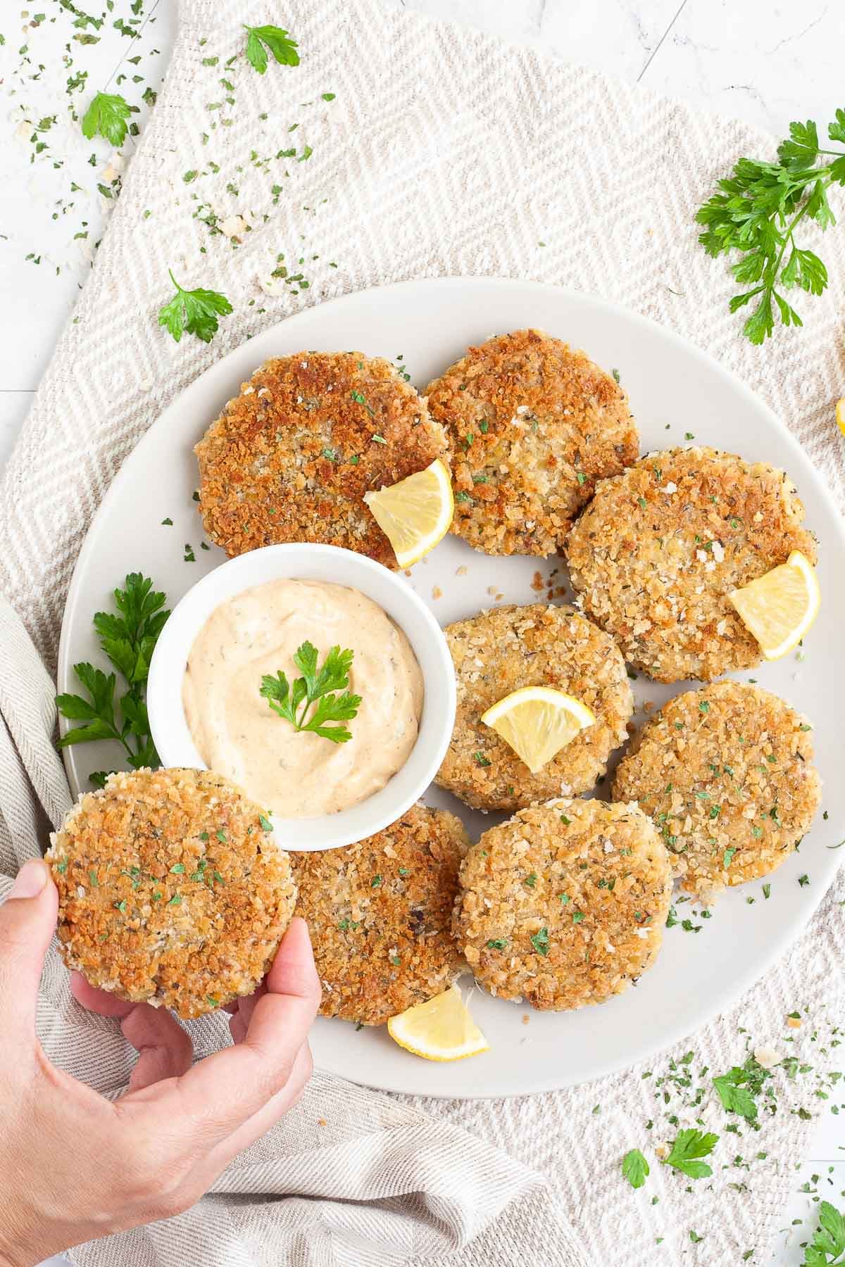 Several round-shaped breadcrumbs covered patties on a large white plate with a light orange sauce in a white bowl. Chopped parsley and lemon wedges are around for decoration. A hand is taking one.
