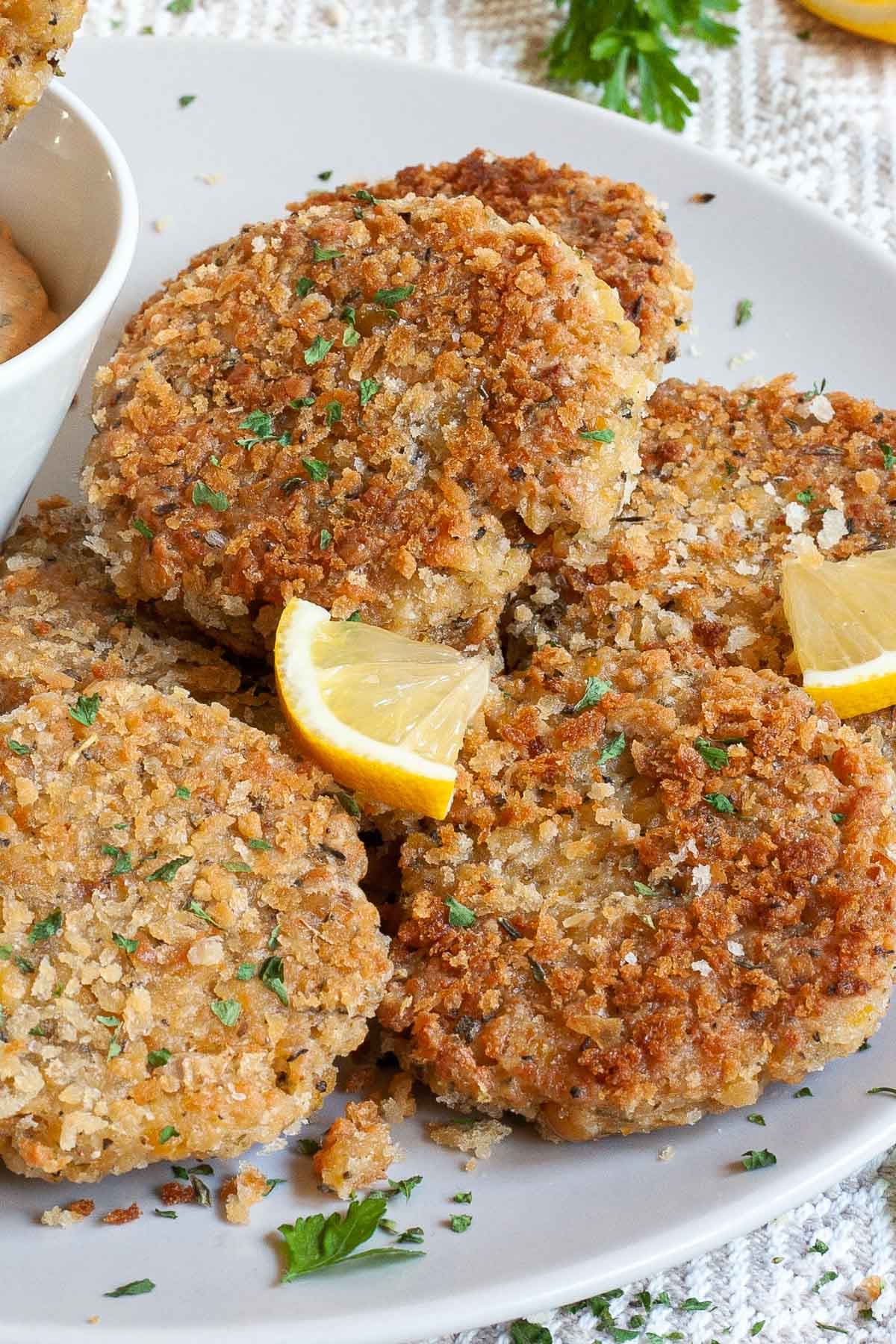 Several round-shaped breadcrumbs covered patties on a large white plate. Chopped parsley and lemon wedges are around for decoration.