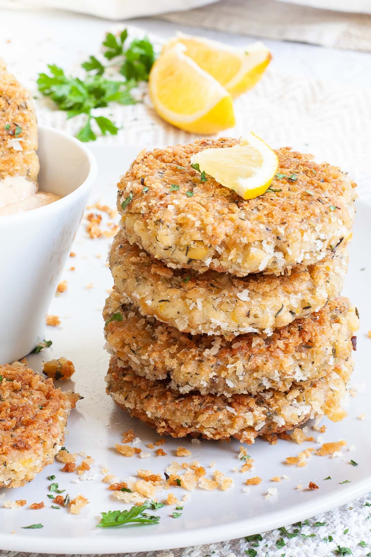 A stack of 4 round-shaped breadcrumbs covered patties on a large white plate with a light orange sauce in a white bowl. Chopped parsley and lemon wedges are around for decoration.