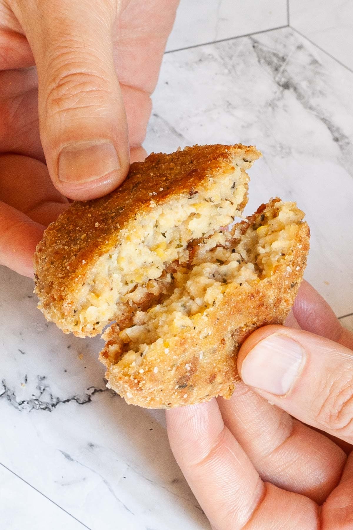 A hand is holding a round breadcrumbs covered patty and tear it in half.