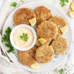 Several round-shaped breadcrumbs covered patties on a large white plate with a light orange sauce in a white bowl. Chopped parsley and lemon wedges are around for decoration.