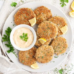 Several round-shaped breadcrumbs covered patties on a large white plate with a light orange sauce in a white bowl. Chopped parsley and lemon wedges are around for decoration.