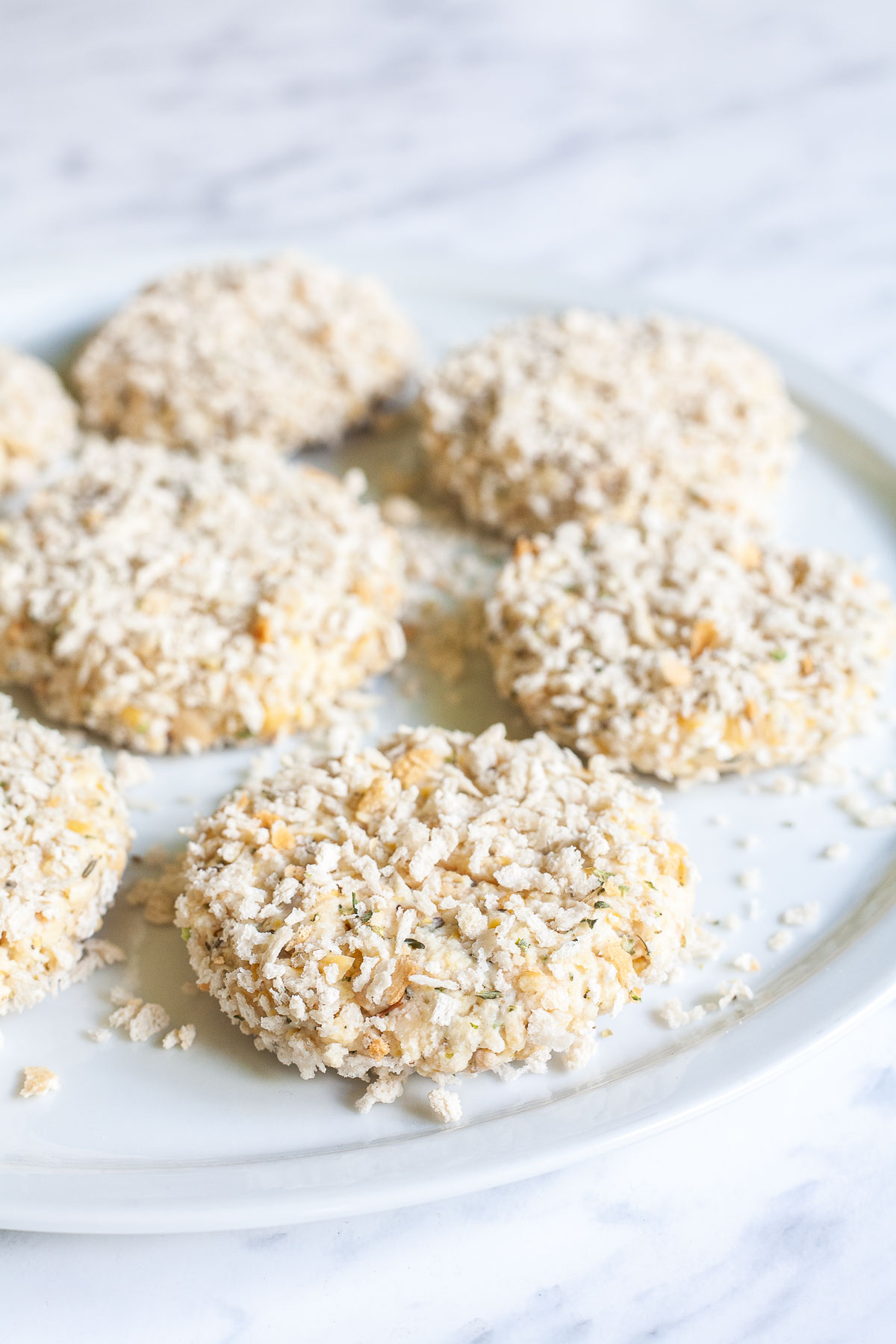 Uncooked round patties covered in breadcrumbs on a white plate.