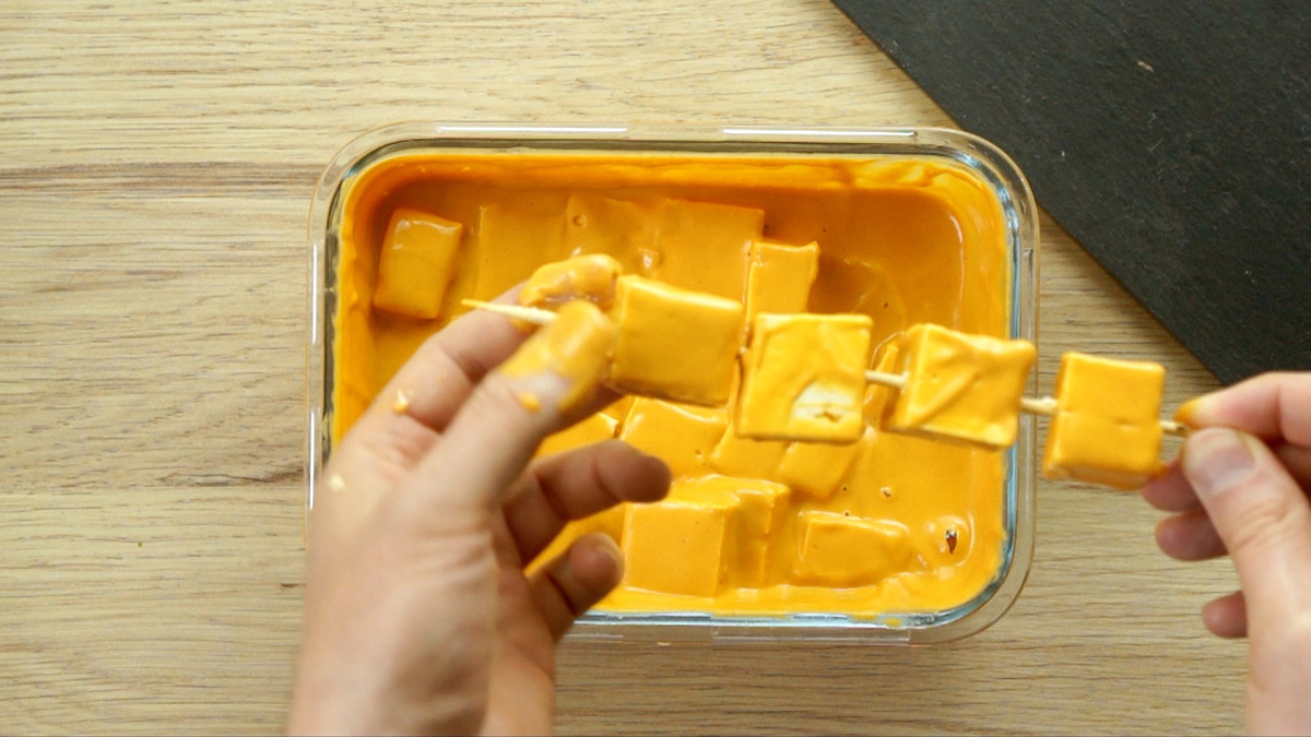 A hand is placing the tofu cubes in thick orange marinade on thin wooden skewers.