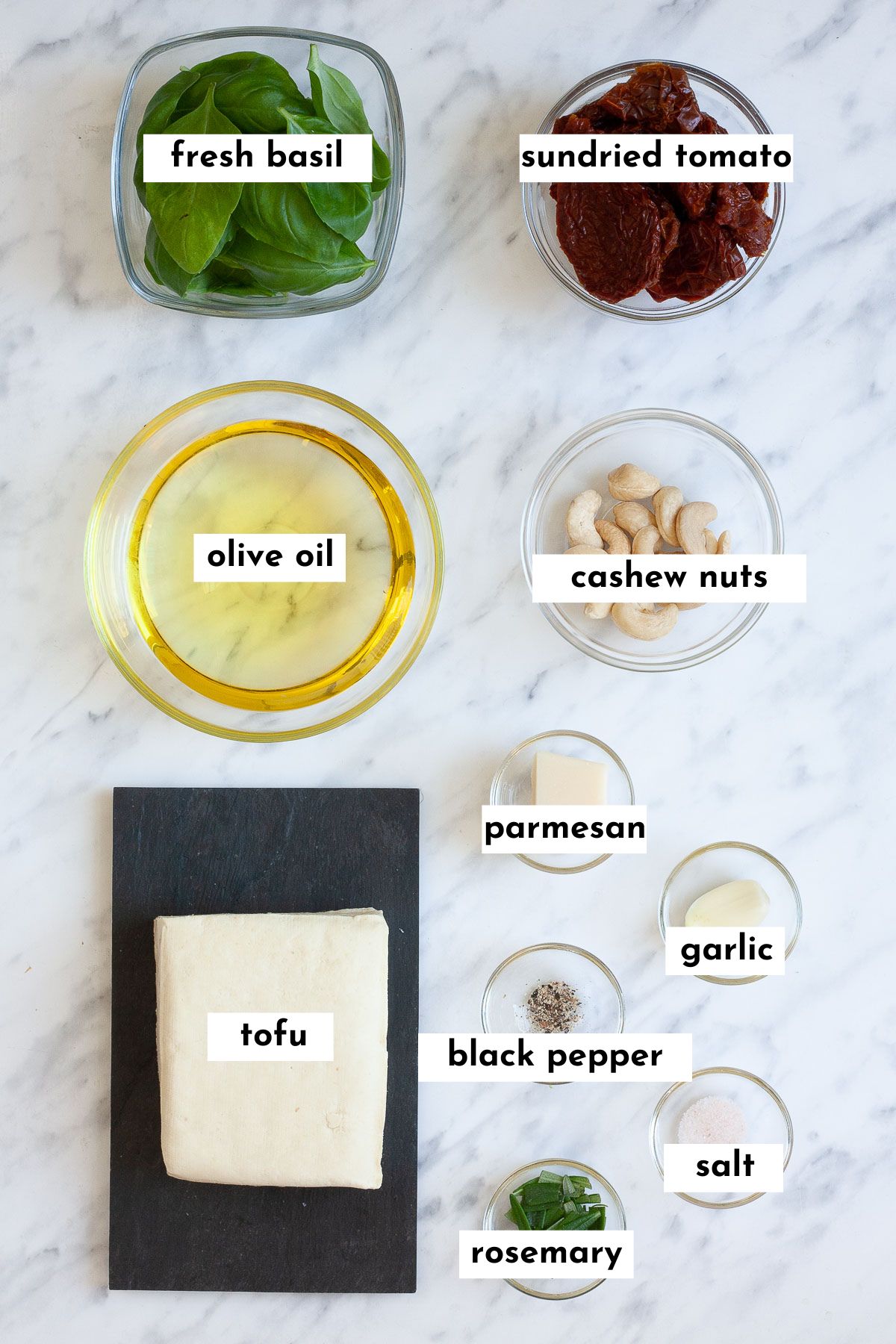 Ingredients of pesto tofu pasta in small glass bowls like basil leaves, oil, sun-dried tomatoes, cashews, garlic clove, a piece of hard cheese, salt, pepper and rosemary leaves. And a block of white tofu is on a black plate. 