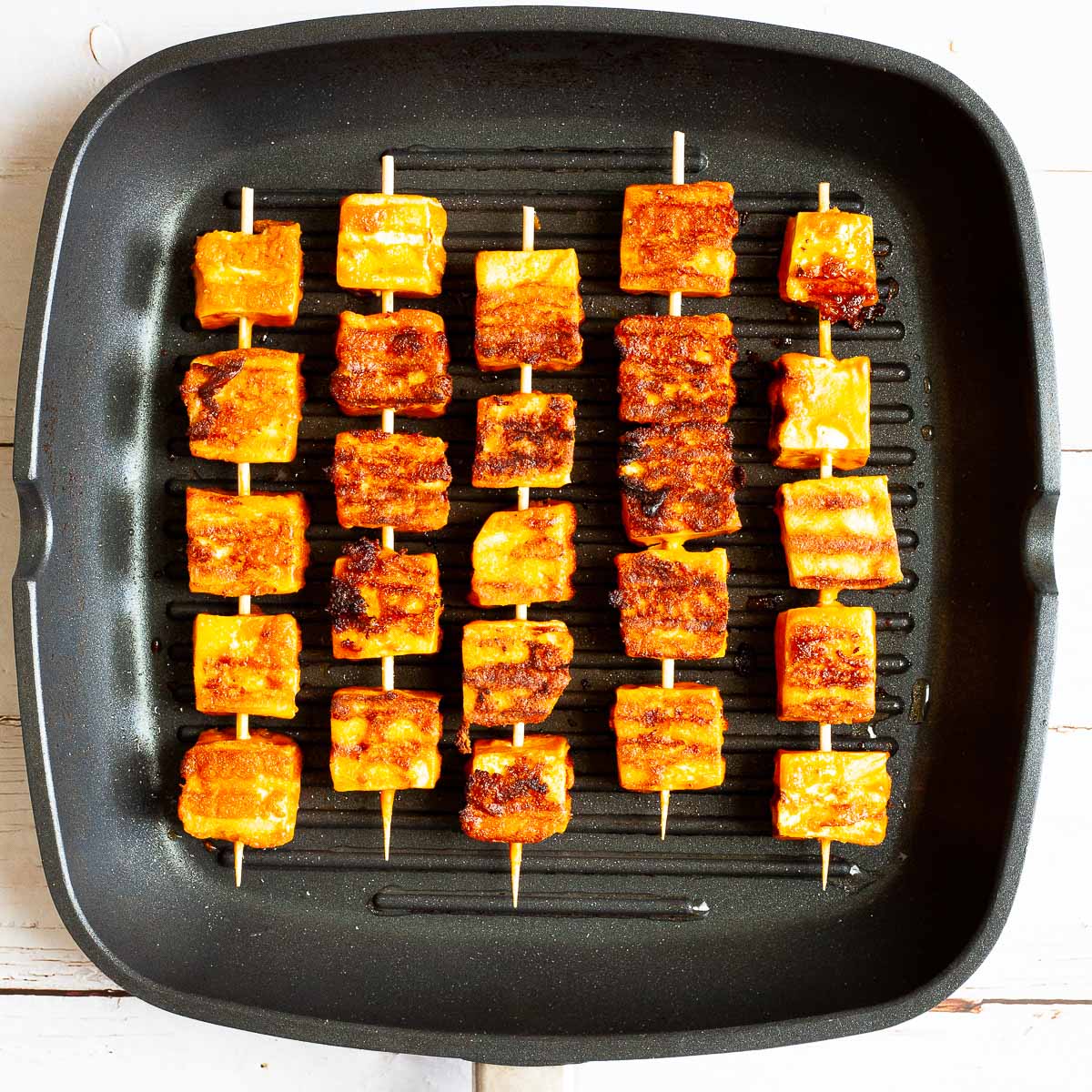 Yellow-brown tofu cubes on 5 wooden skewers are placed lengthwise in a grill pan.