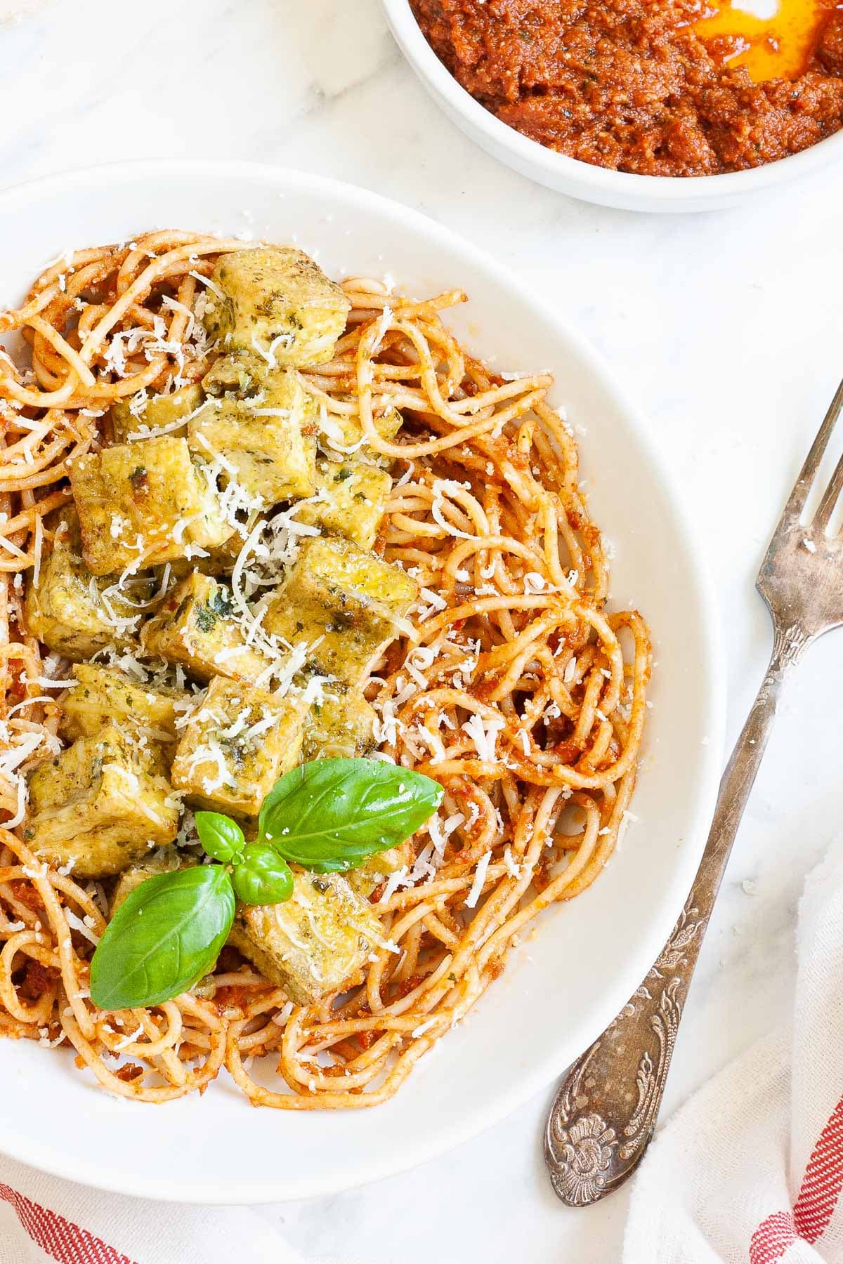 Large white plate with red pesto spaghetti topped with green-yellow tofu cubes, shredded cheese and basil leaves. There is a small bowl with leftover red pesto in the corner.
