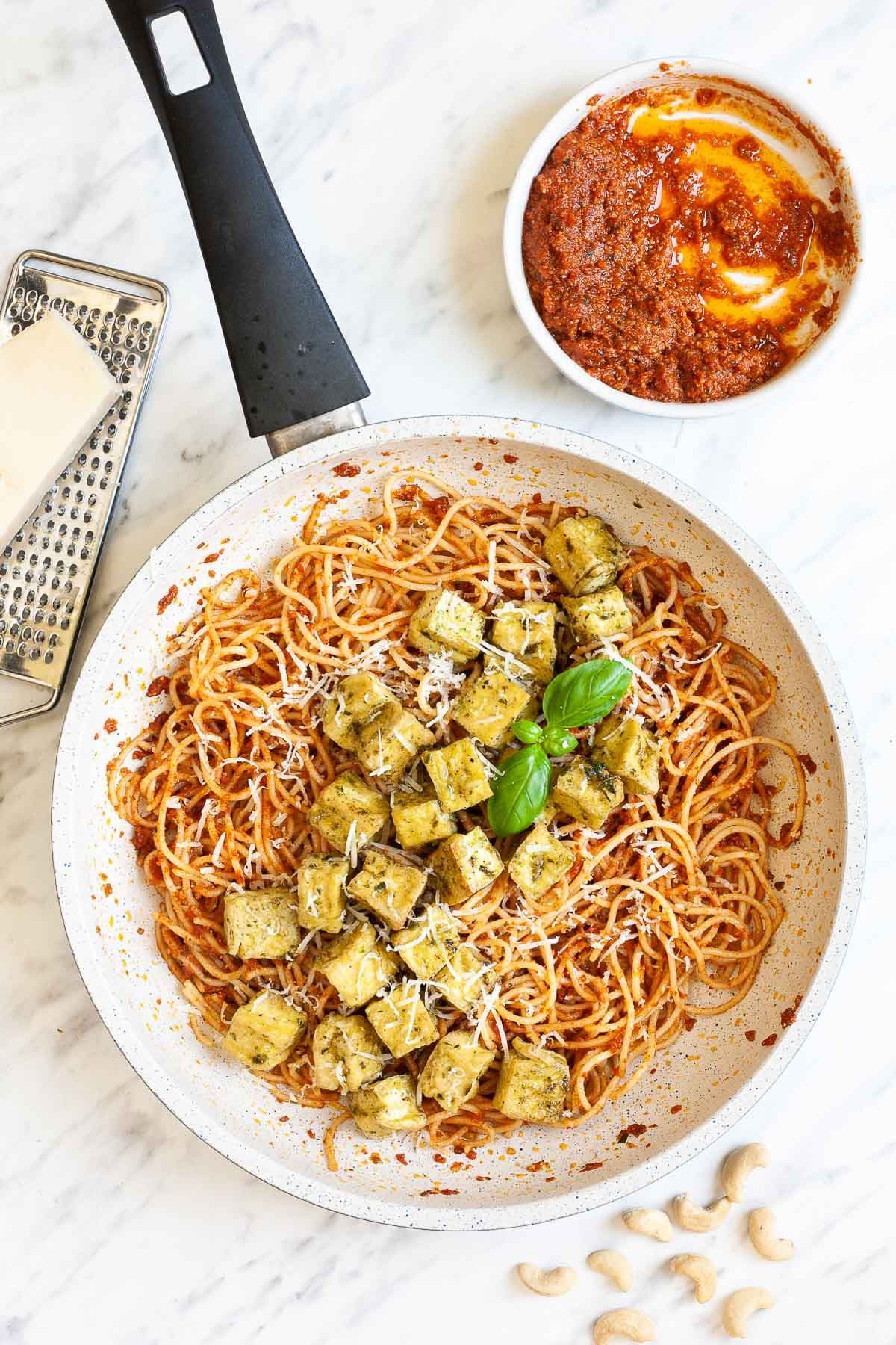 Large white frying pan with red pesto spaghetti topped with green-yellow tofu cubes, shredded cheese and basil leaves. There is a small bowl with leftover red pesto in the corner and parmesan cheese on top of a grater.