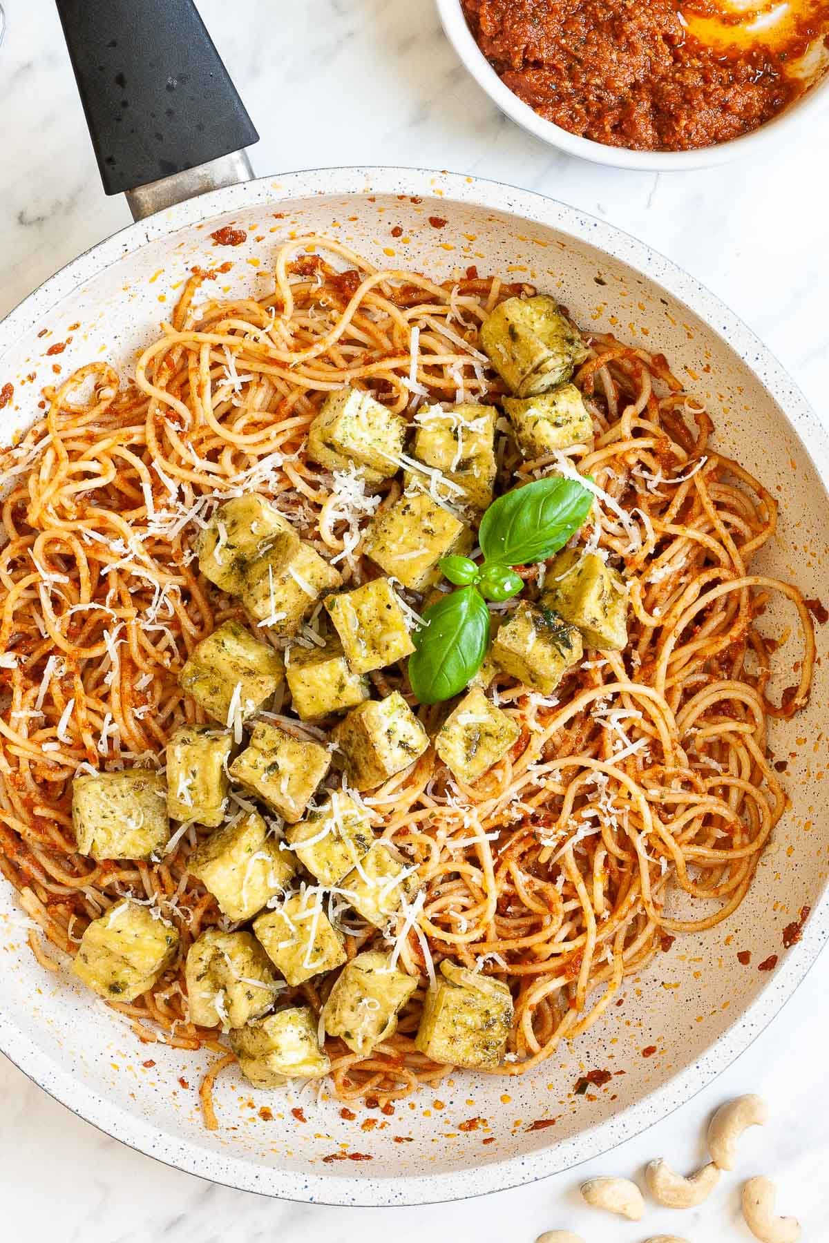 Large white frying pan with red pesto spaghetti topped with green-yellow tofu cubes, shredded cheese and basil leaves.
