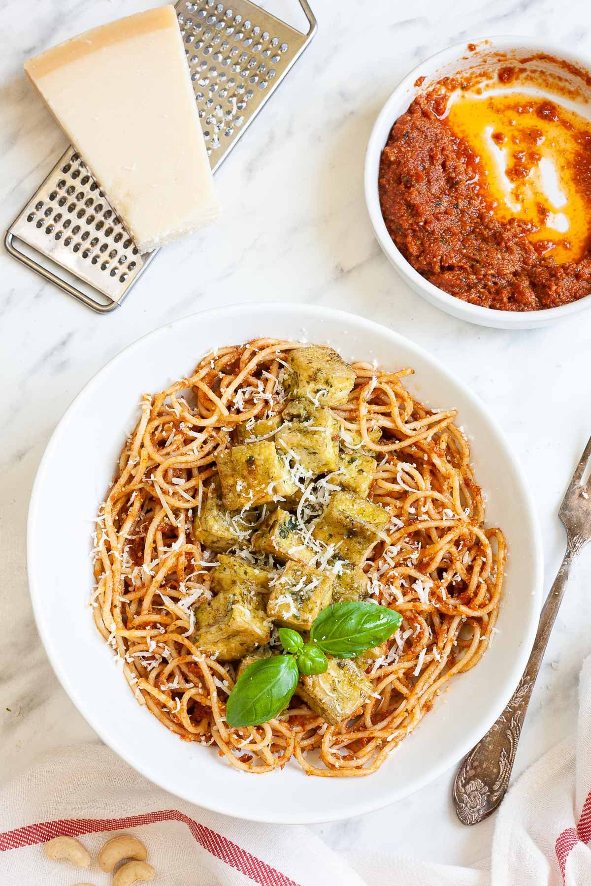 Large white plate with red pesto spaghetti topped with green-yellow tofu cubes, shredded cheese and basil leaves. There is a small bowl with leftover red pesto in the corner and parmesan cheese on top of a grater.
