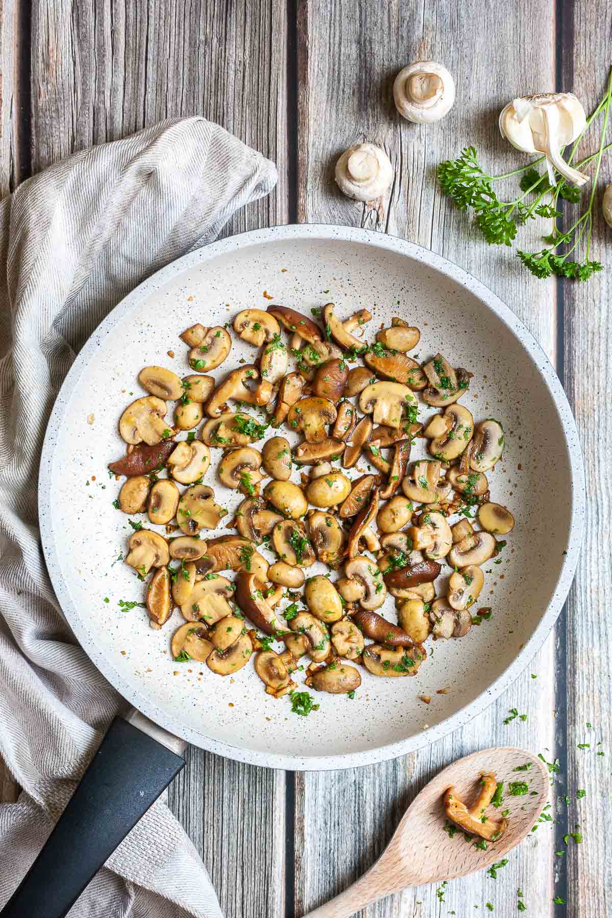 Golden brown sliced button and shiitake mushrooms in a frying pan topped with freshly chopped greens. A wooden spoon is next to it as well as some raw ingredients.