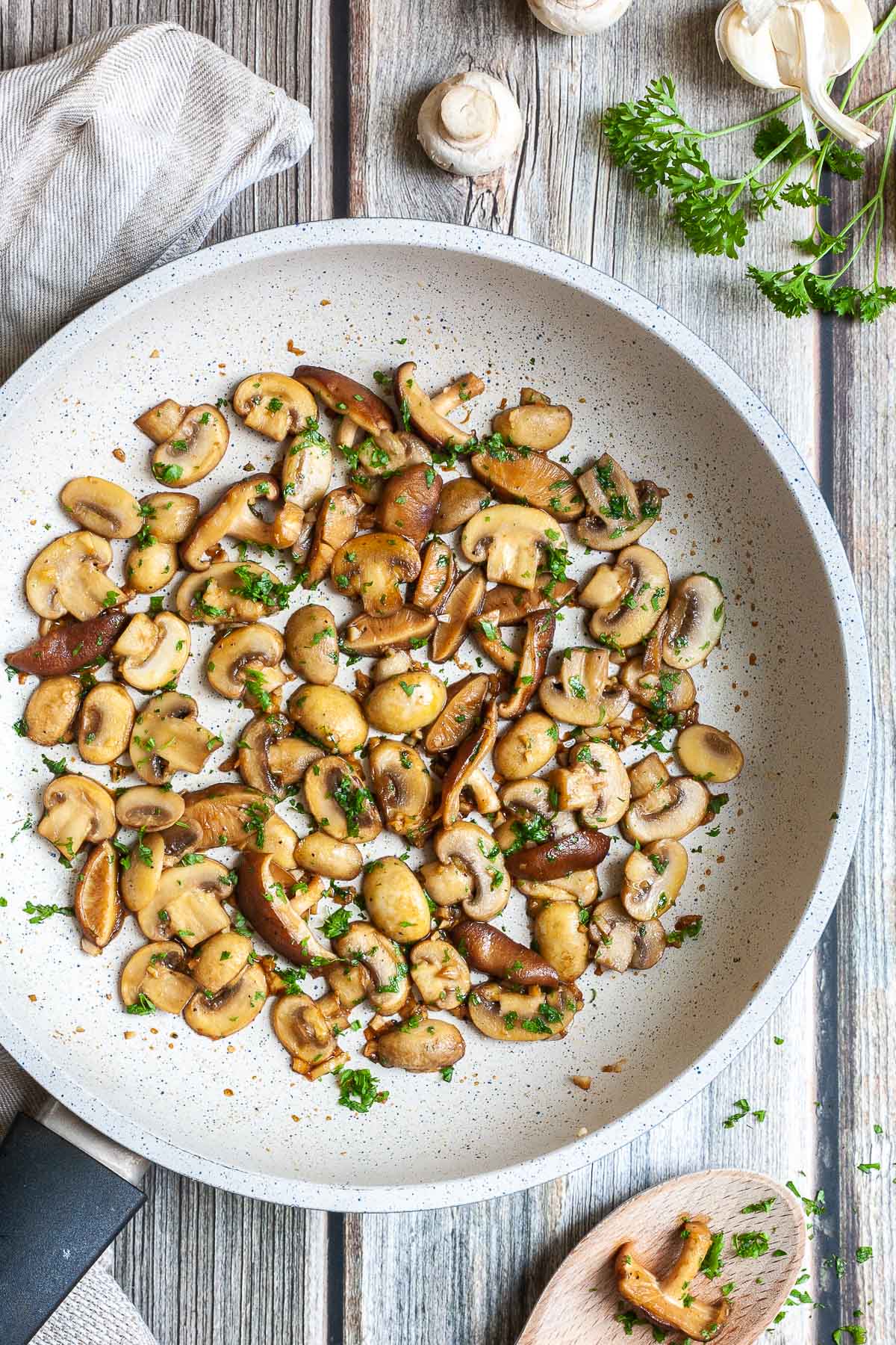 Golden brown sliced button and shiitake mushrooms in a frying pan topped with freshly chopped greens. A wooden spoon is next to it as well as some raw ingredients. 