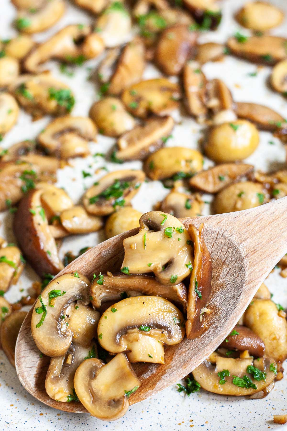 Golden brown sliced button and shiitake mushrooms in a frying pan topped with freshly chopped greens. A wooden spoon is taking some from the middle.