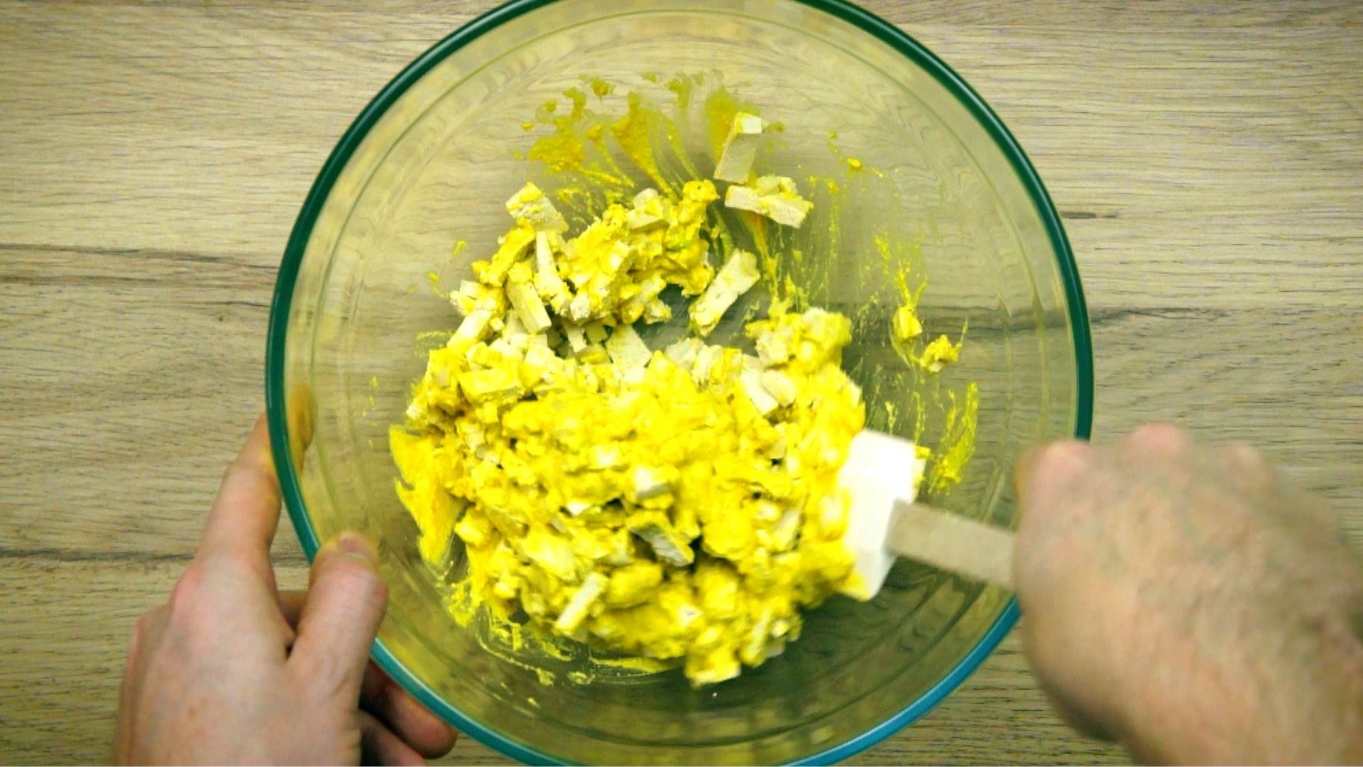A glass bowl with white tofu cubes mixed with a yellow sauce by a hand holding a silicone spatula. 