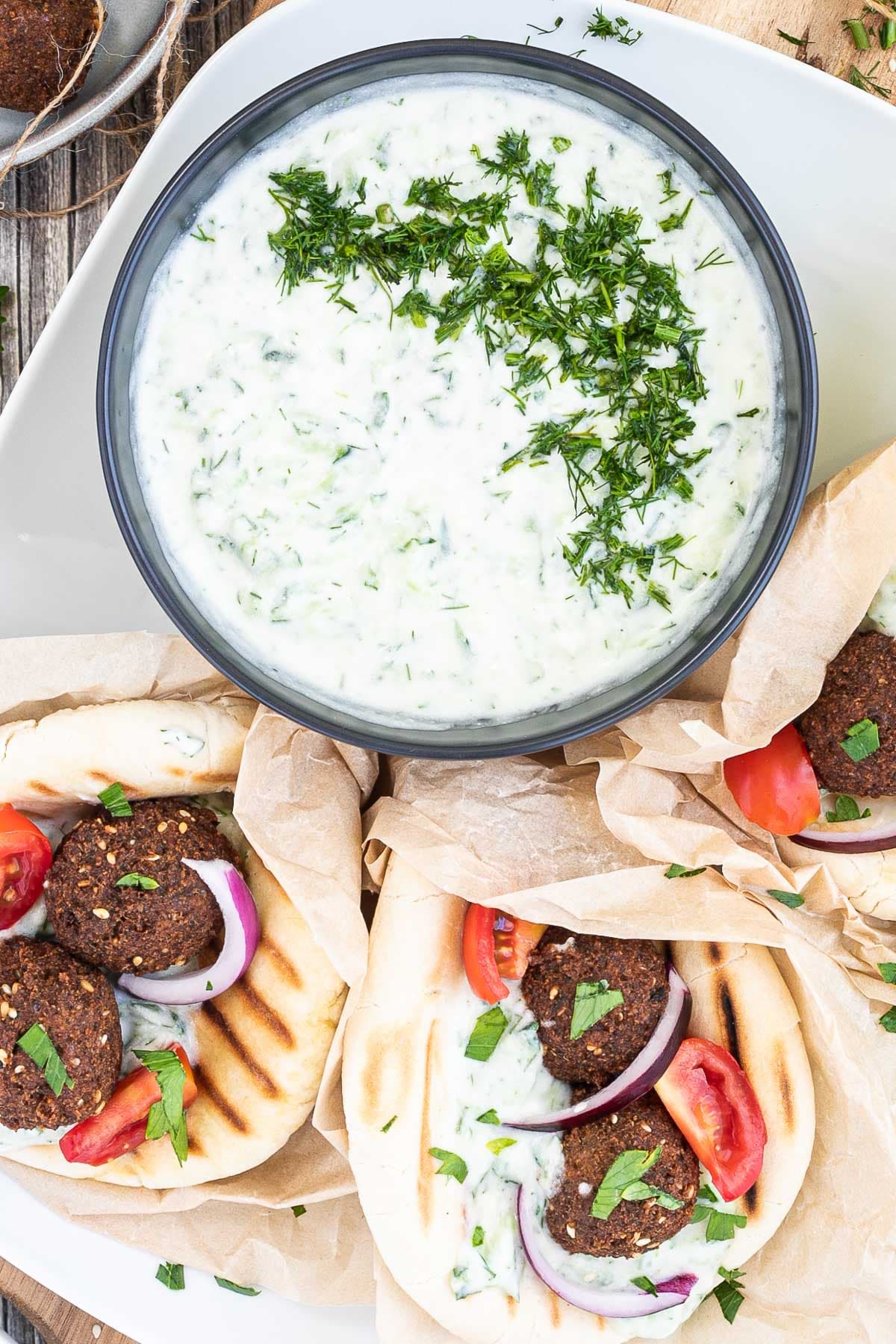 3 pitas folded and stuffed with 2 brown falafel balls, white grated cucumber salad, tomato and purple onion slices. A black bowl of white grated cucumber sauce is placed next to them.