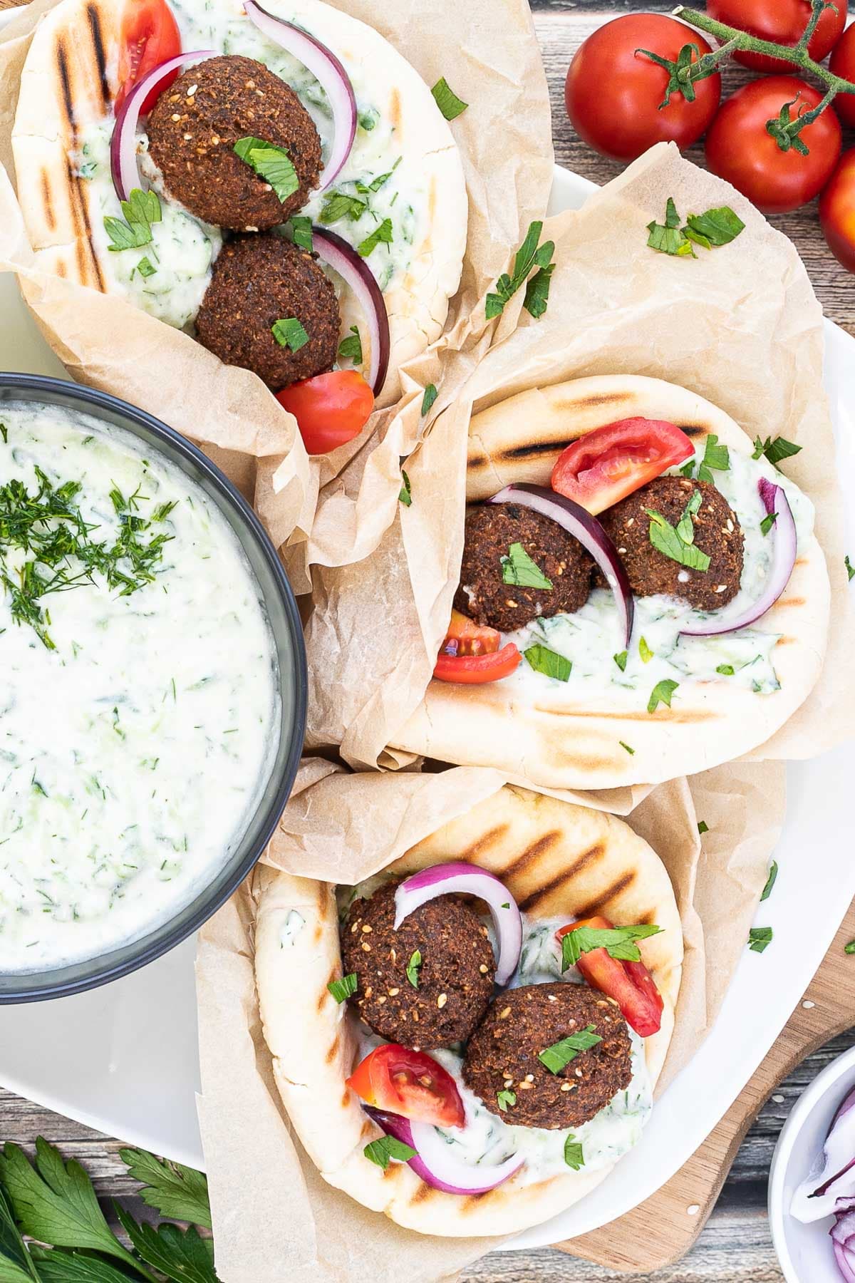 3 pitas folded and stuffed with 2 brown falafel balls, white grated cucumber salad, tomato and purple onion slices. A black bowl of white grated cucumber sauce is placed next to them.