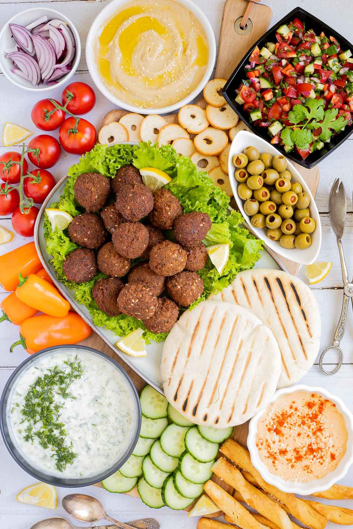 Lot of items in a white board including falafel balls on top of lettuce leaves, charcoaled pita breads, grated cucumber in yoghurt sauce, orange hummus, olives, chopped green red salad, light brown sauce, cherry tomatoes, orange peppers, sliced cucumber, breadsticks and crackers. 