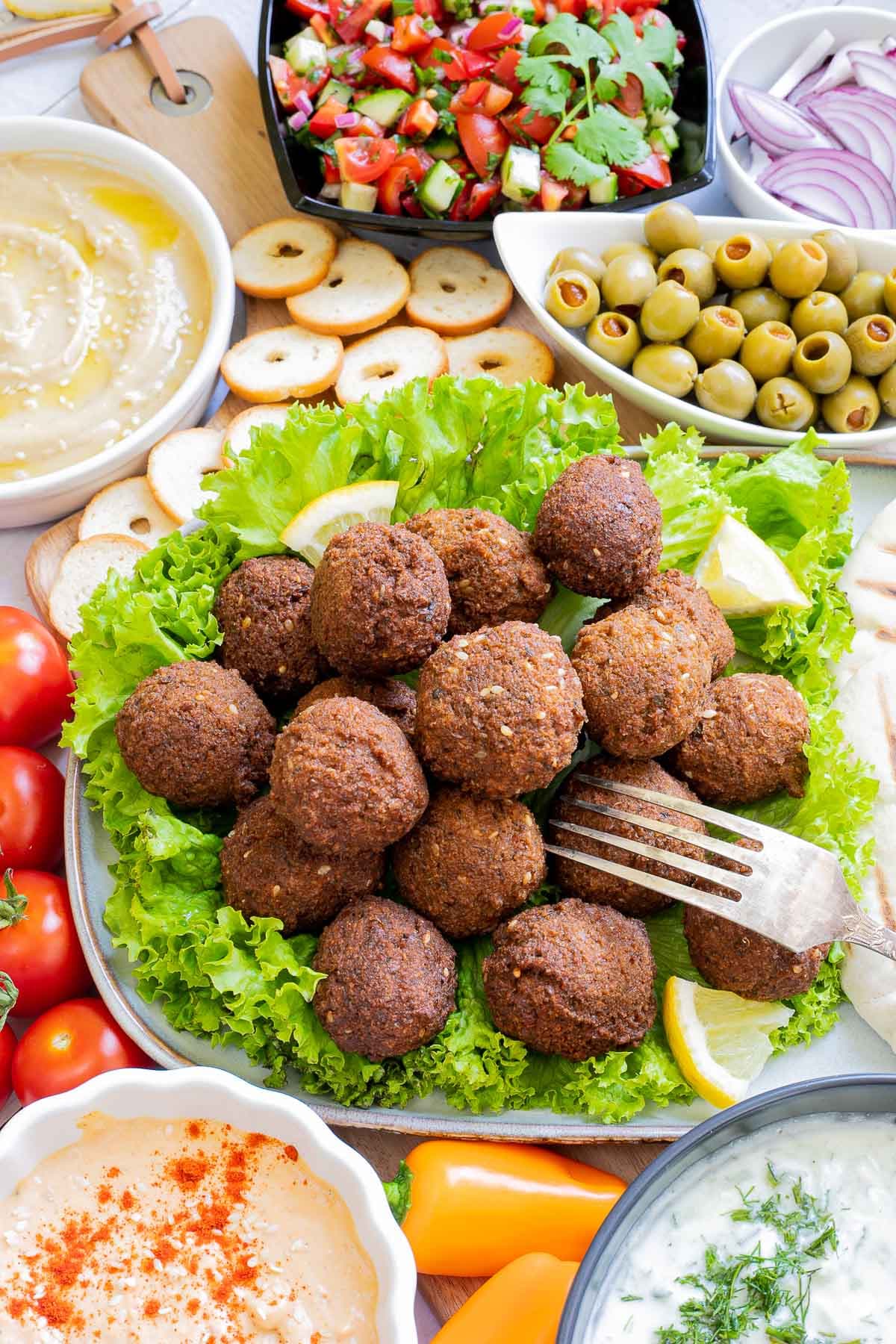 Lot of items in a white board including falafel balls on top of lettuce leaves, charcoaled pita breads, grated cucumber in yoghurt sauce, orange hummus, olives, chopped green red salad, light brown sauce, cherry tomatoes, orange peppers, sliced cucumber, breadsticks and crackers.