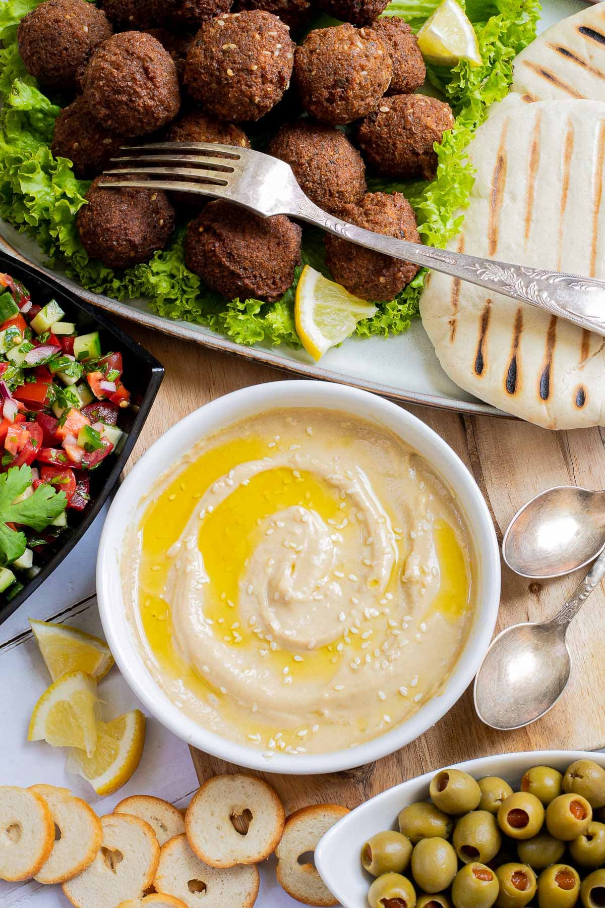 Light brown sauce in a white shallow bowl sprinkled with white sesame seeds. Falafel balls, pita breads, and chopped red green veggie salad is on the side.