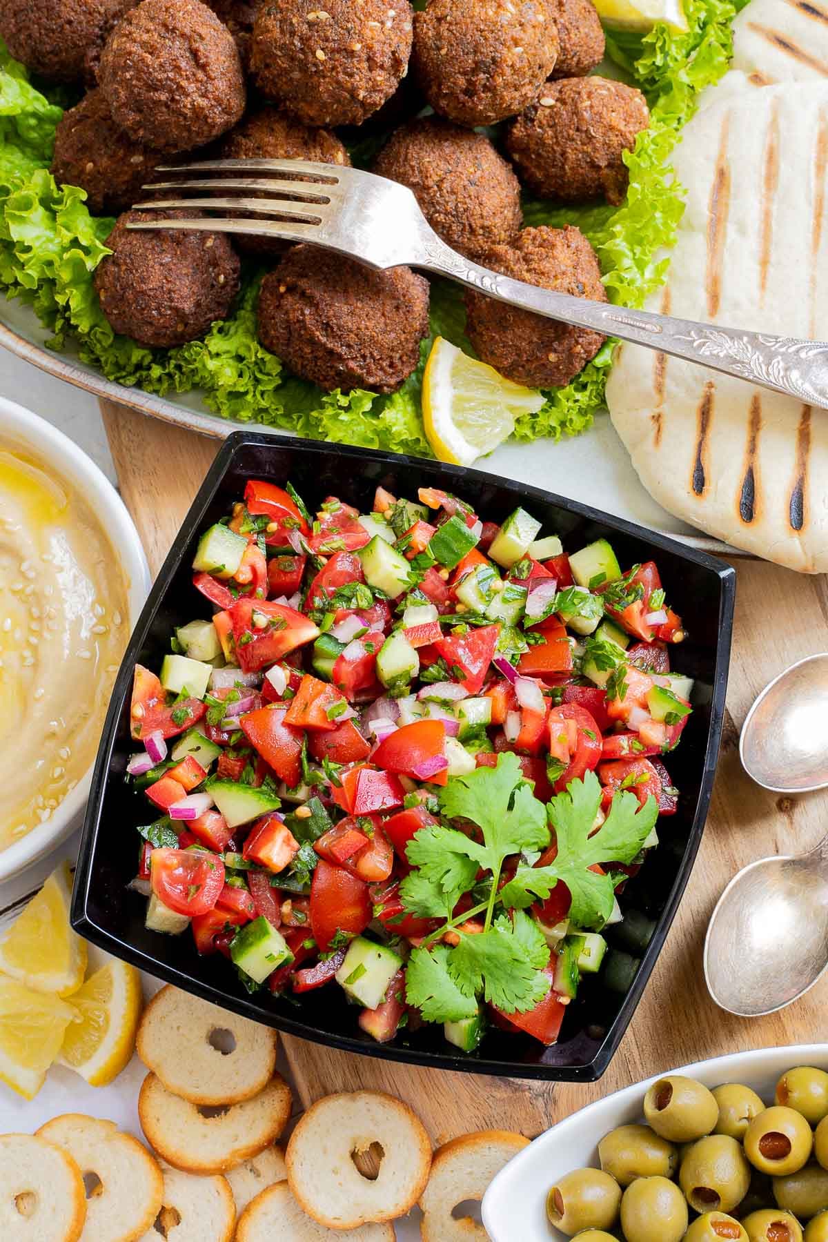 Chopped red green veggie salad in a black bowl. Falafel balls, pita breads, and light brown sauce in shallow white bowl sprinkled with sesame seeds.