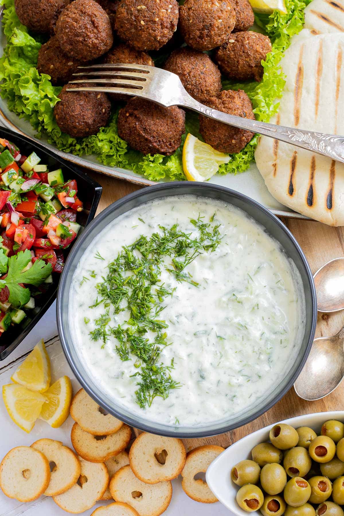 Grated cucumber in white yogurt sauce in a black bowl. Falafel balls, pita breads, green olives, and chopped red green veggie salad is on the side.