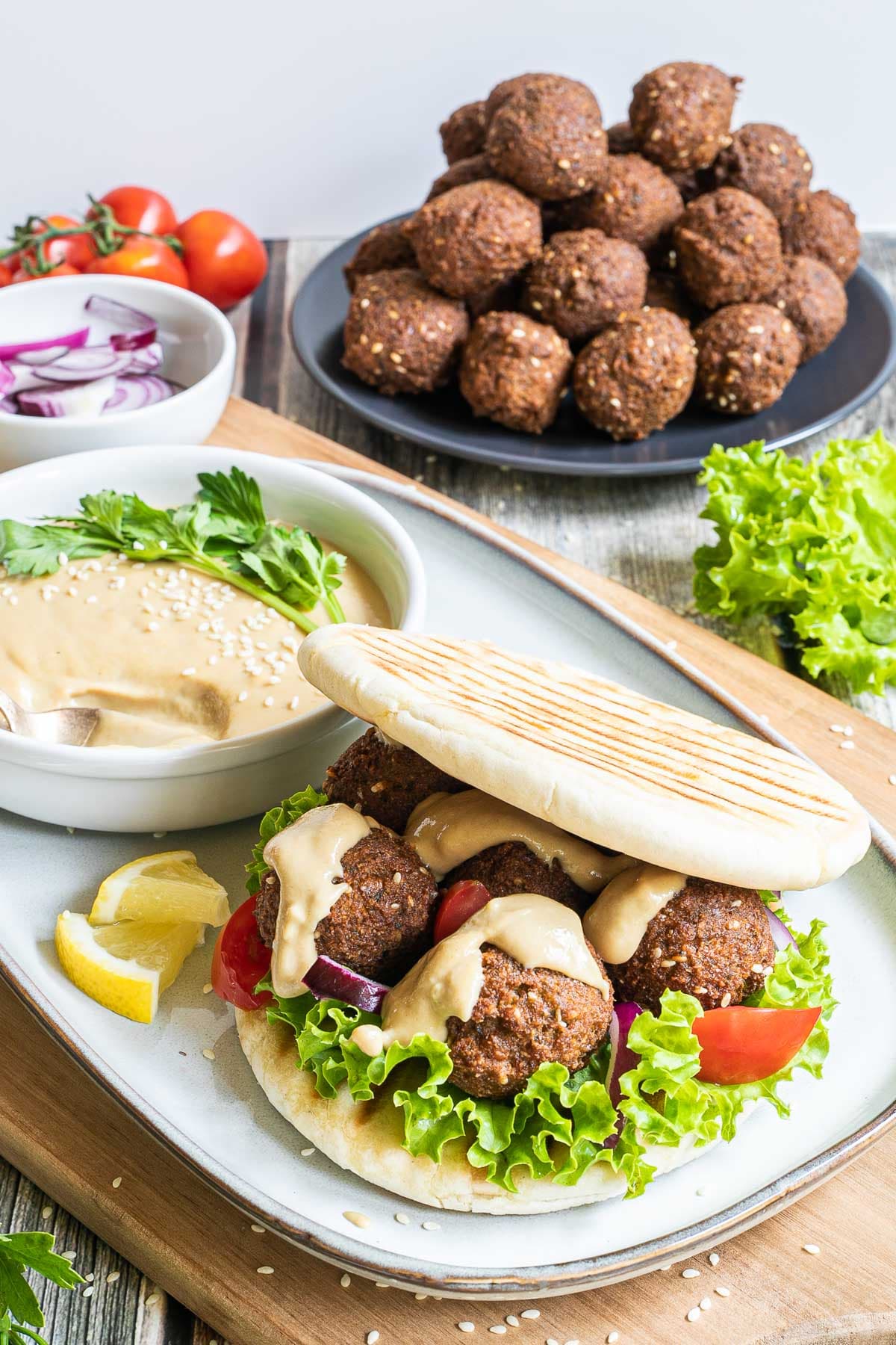 Two pita breads stuffed with 4 dark brown falafel balls, drizzled with a light brown sauce with sliced purple onion, tomato pieces and lettuce. All elements of the sandwich is around it on different plates.