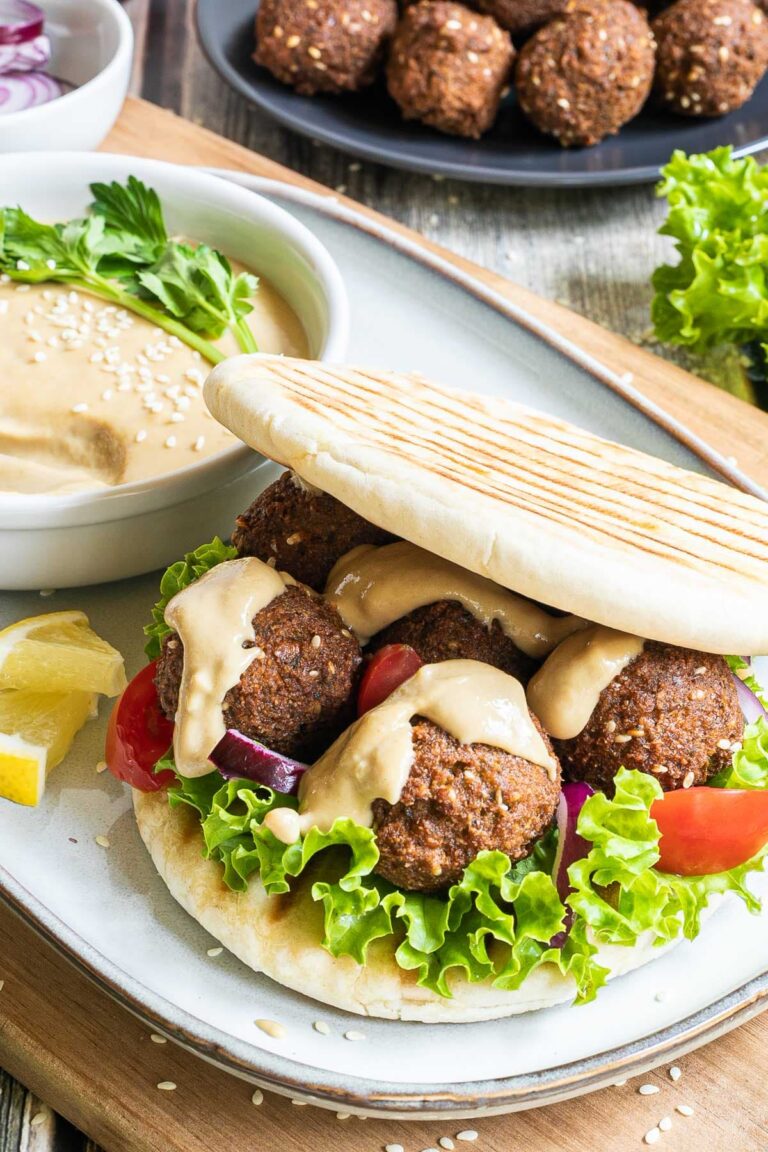 Two pita breads stuffed with 4 dark brown falafel balls, drizzled with a light brown sauce with sliced purple onion, tomato pieces and lettuce. All elements of the sandwich is around it on different plates.