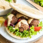 Two pita breads stuffed with 4 dark brown falafel balls, drizzled with a light brown sauce with sliced purple onion, tomato pieces and lettuce.