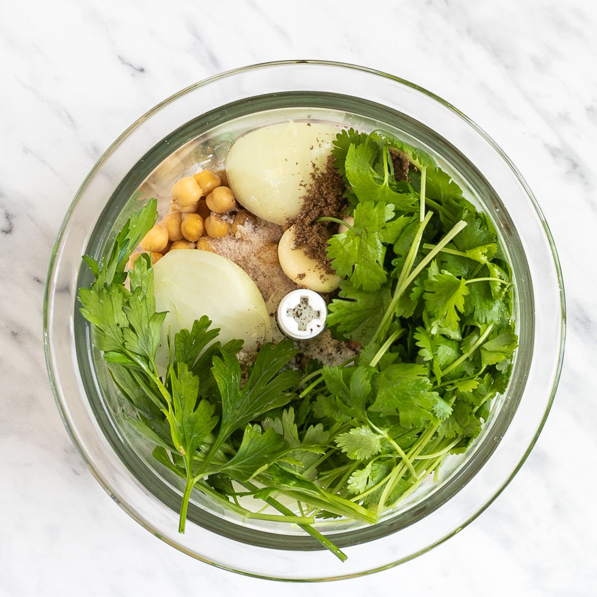 A glass food processor container from above first with raw ingredients like green herbs, chickpeas and onion