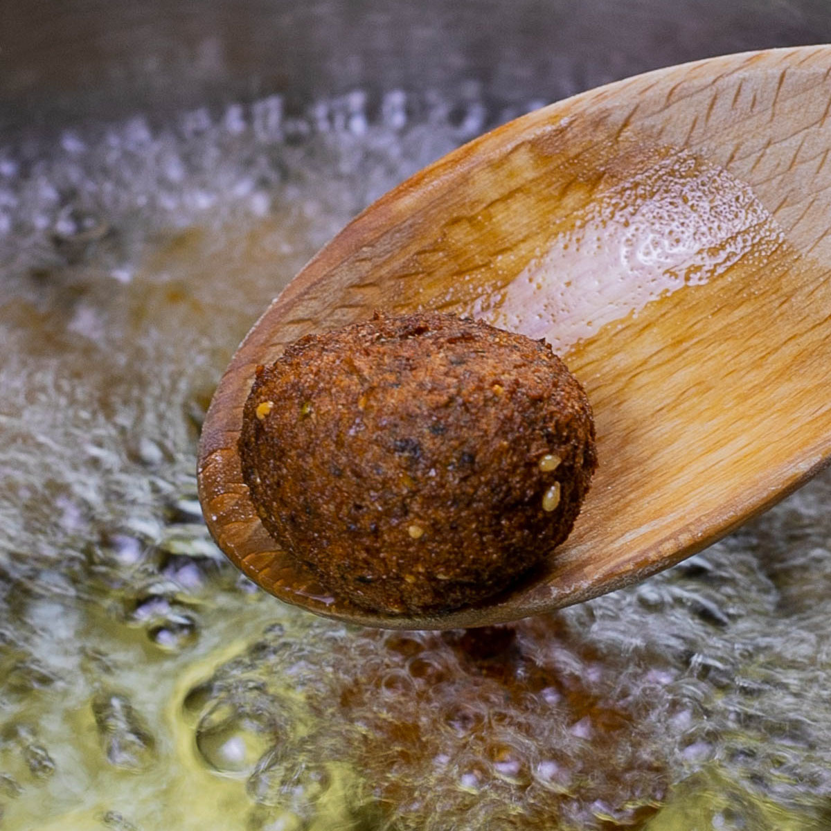 A wooden spoon with one dark brown crispy falafel ball just above a pan of boiling oil.