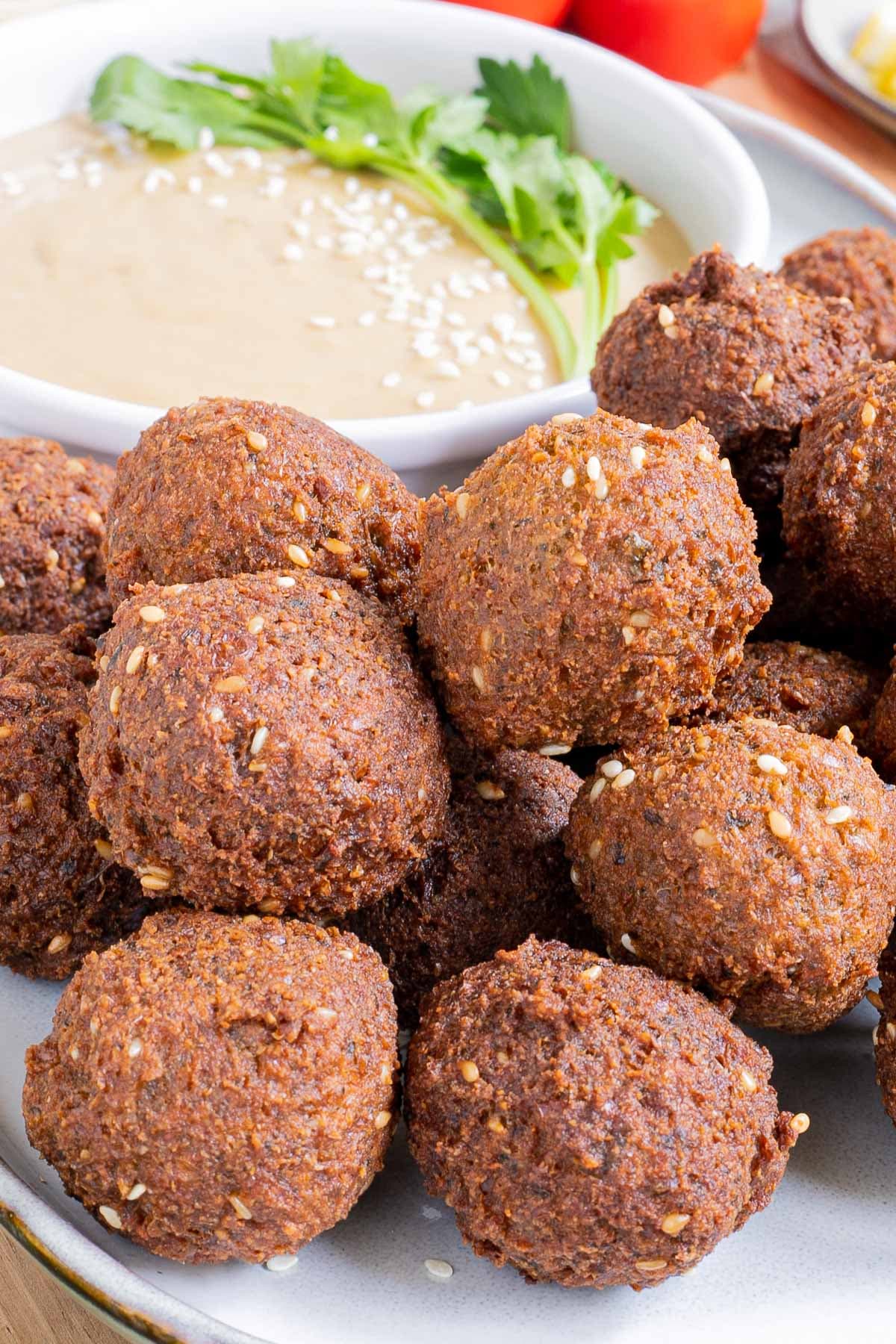 Falafel balls are served on a large plate. A light brown sauce in a white shallow bowl sprinkled with white sesame seeds and topped with a twig of parsley is next to them..