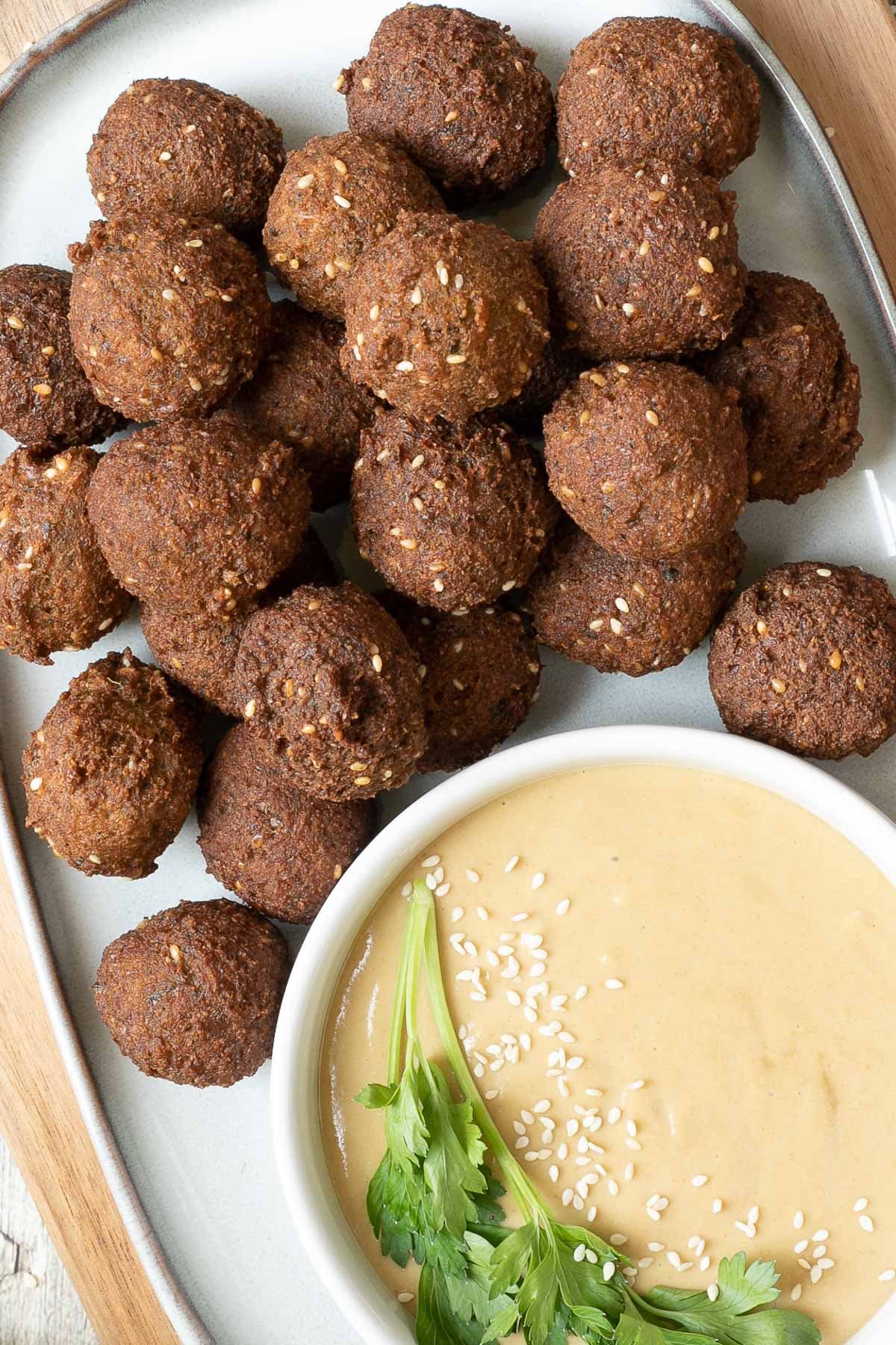 Falafel balls are served on a large plate. A light brown sauce in a white shallow bowl sprinkled with white sesame seeds and topped with a twig of parsley is next to them..