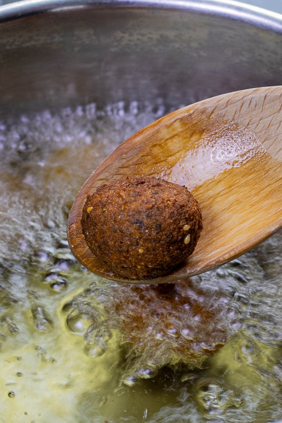 A wooden spoon with one dark brown crispy falafel ball just above a pan of boiling oil.