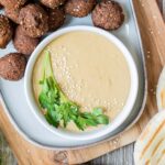 Light brown sauce in a white shallow bowl sprinkled with white sesame seeds and topped with a twig of parsley. Falafel balls and pite breads are next to it.