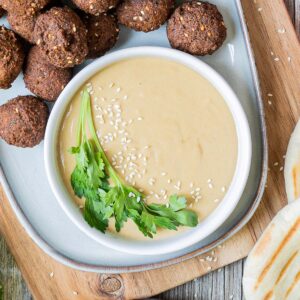 Light brown sauce in a white shallow bowl sprinkled with white sesame seeds and topped with a twig of parsley. Falafel balls and pite breads are next to it.