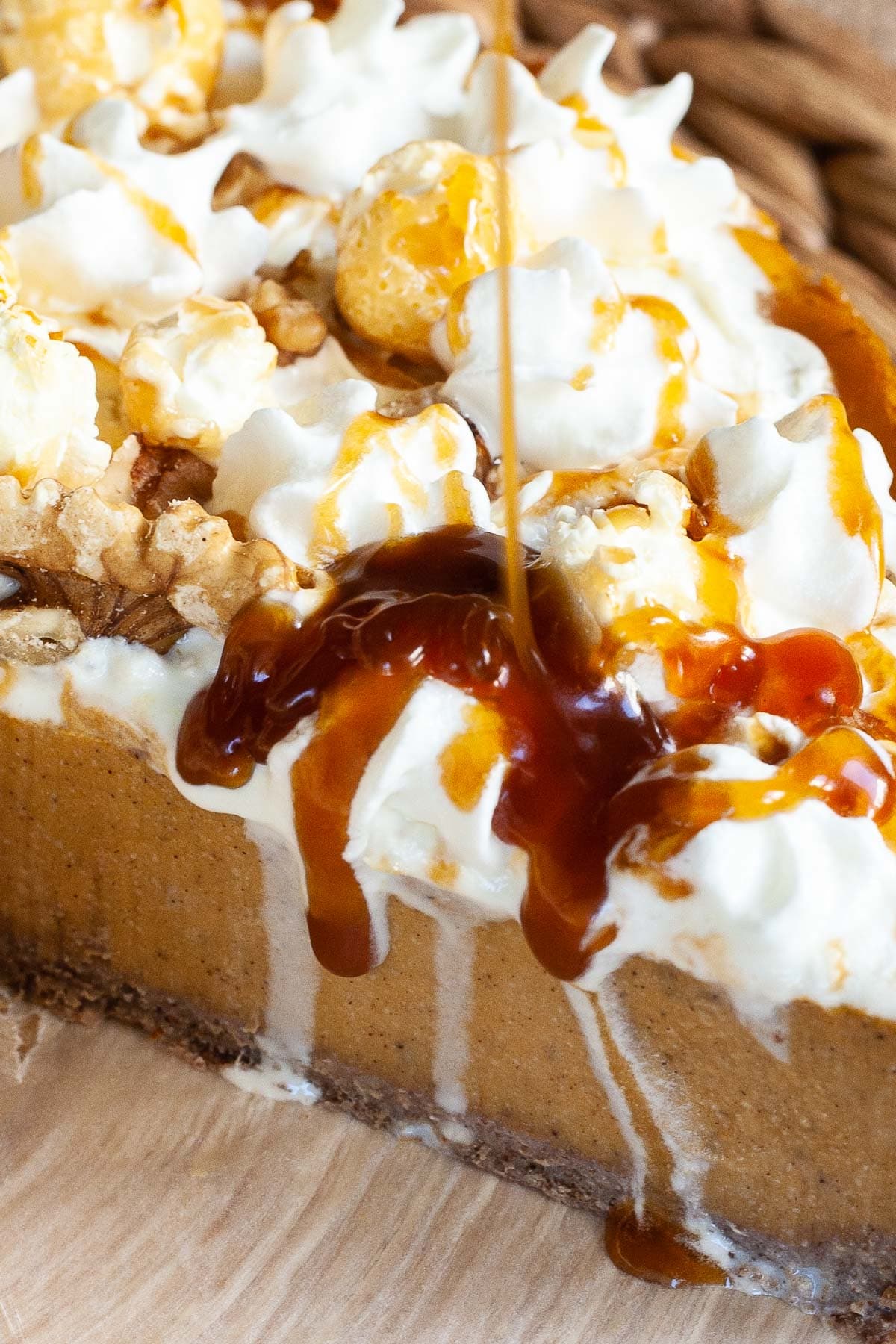 Part of an amber brown cheesecake topped with whipped cream, caramel sauce and chopped nuts. Caramel sauce is flowing right on the top.