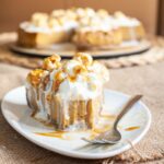 One slice of amber brown cheesecake with whipped cream, caramel sauce and chopped nuts on top is served on a white plate. One bite is missing from the front. The whole cake is in the background.