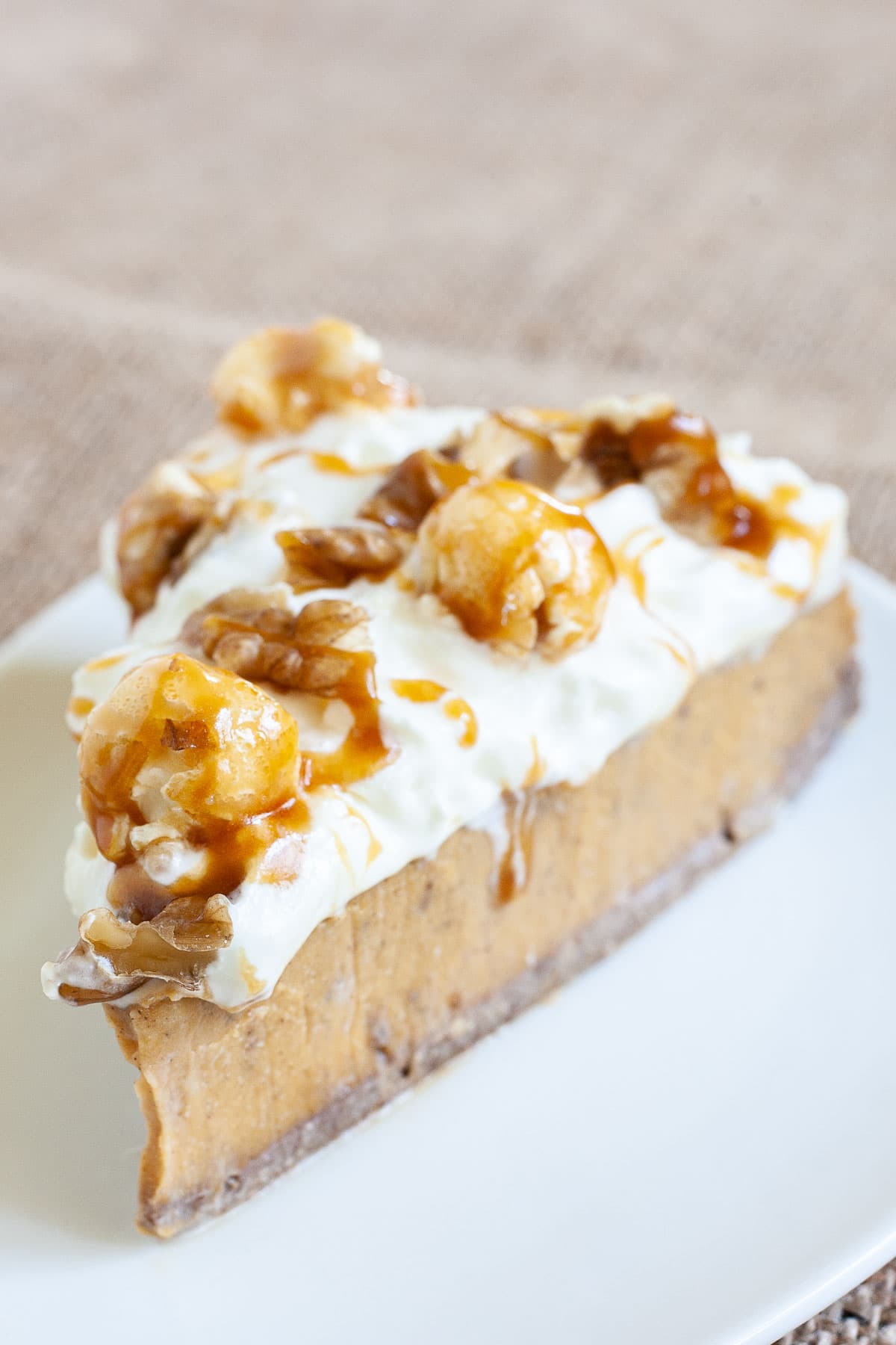One slice of amber brown cheesecake with whipped cream, caramel sauce and chopped nuts on top is served on a white plate.
