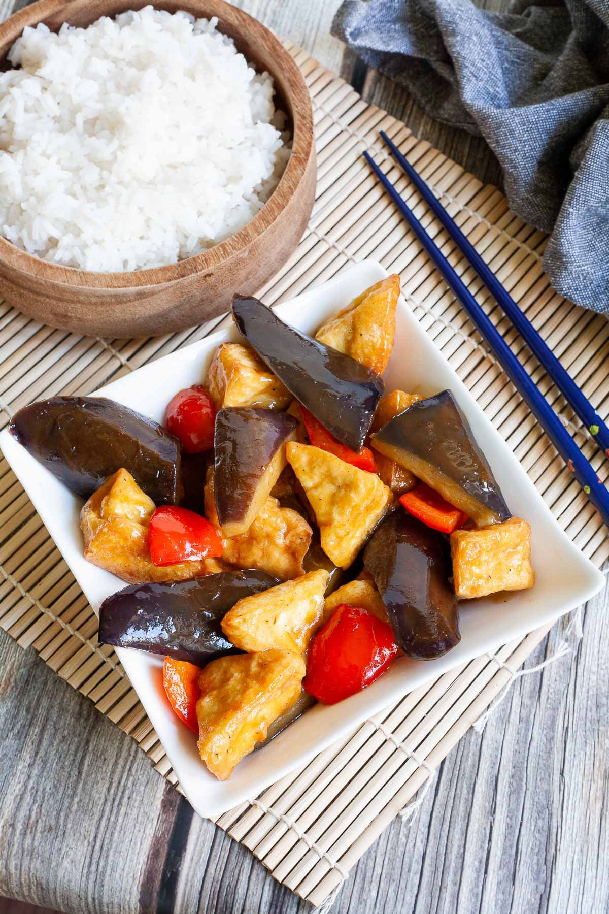 White rectangular plate full of large purple eggplant chunks, puffy brown glazed tofu and red peppers. Rice is served in a wooden bowl next to it.