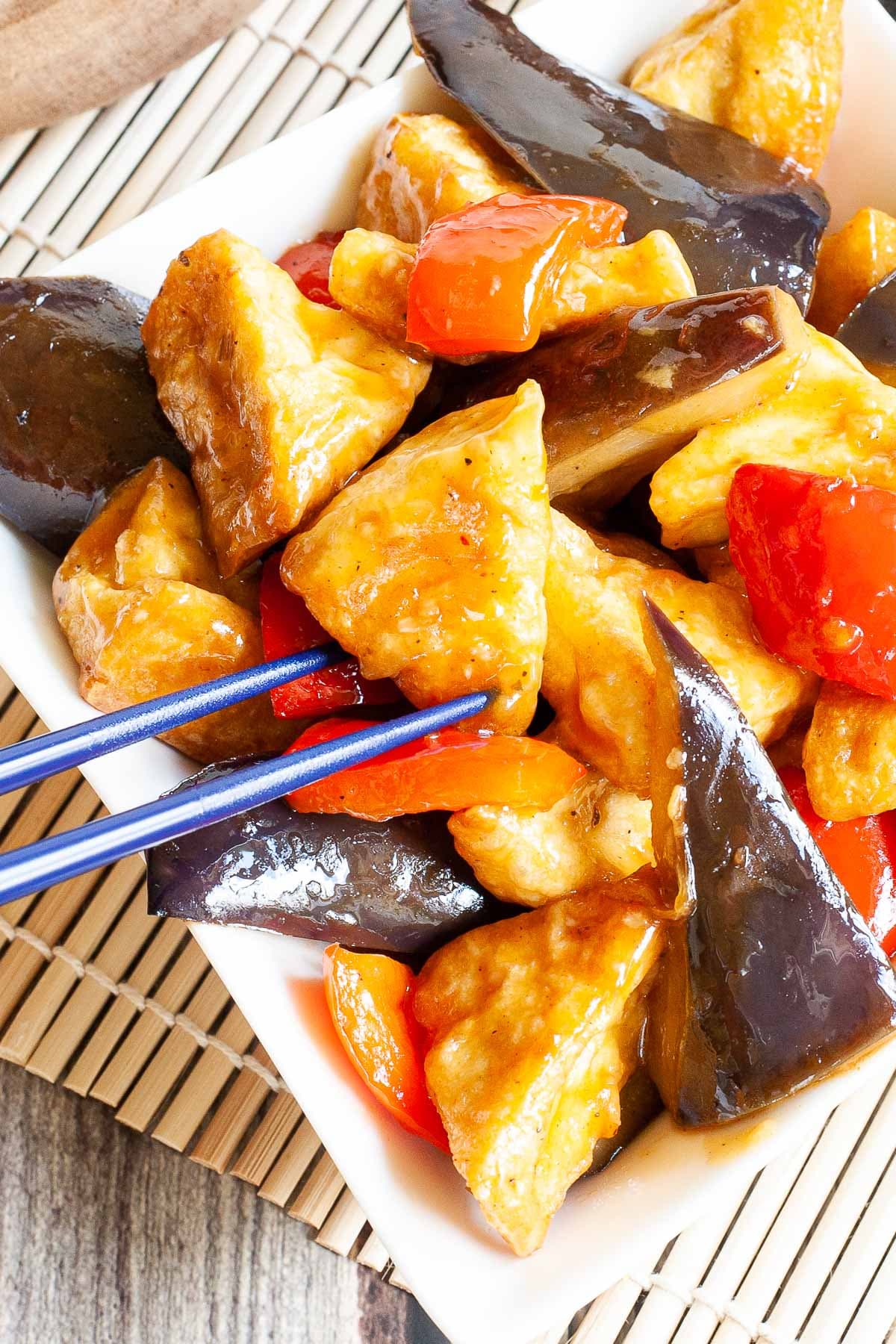 White rectangular plate full of large purple eggplant chunks, puffy brown glazed tofu and red peppers. Blue chopsticks are taking one tofu piece.