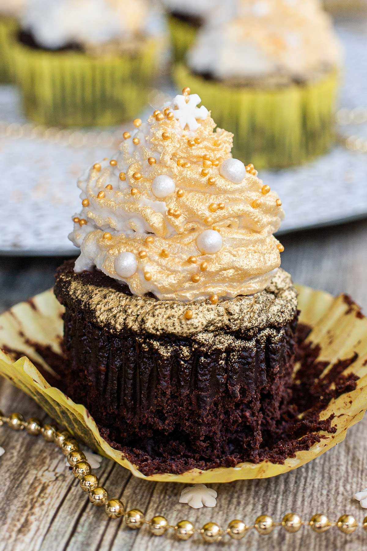 A dark brownie cupcake in yellow unwrapped cupcake liner topped with a white frosting sprayed with gold and sprinkled with gold and white pearl sprinkles and a snowflake one on top. Other cupcakes at the back.